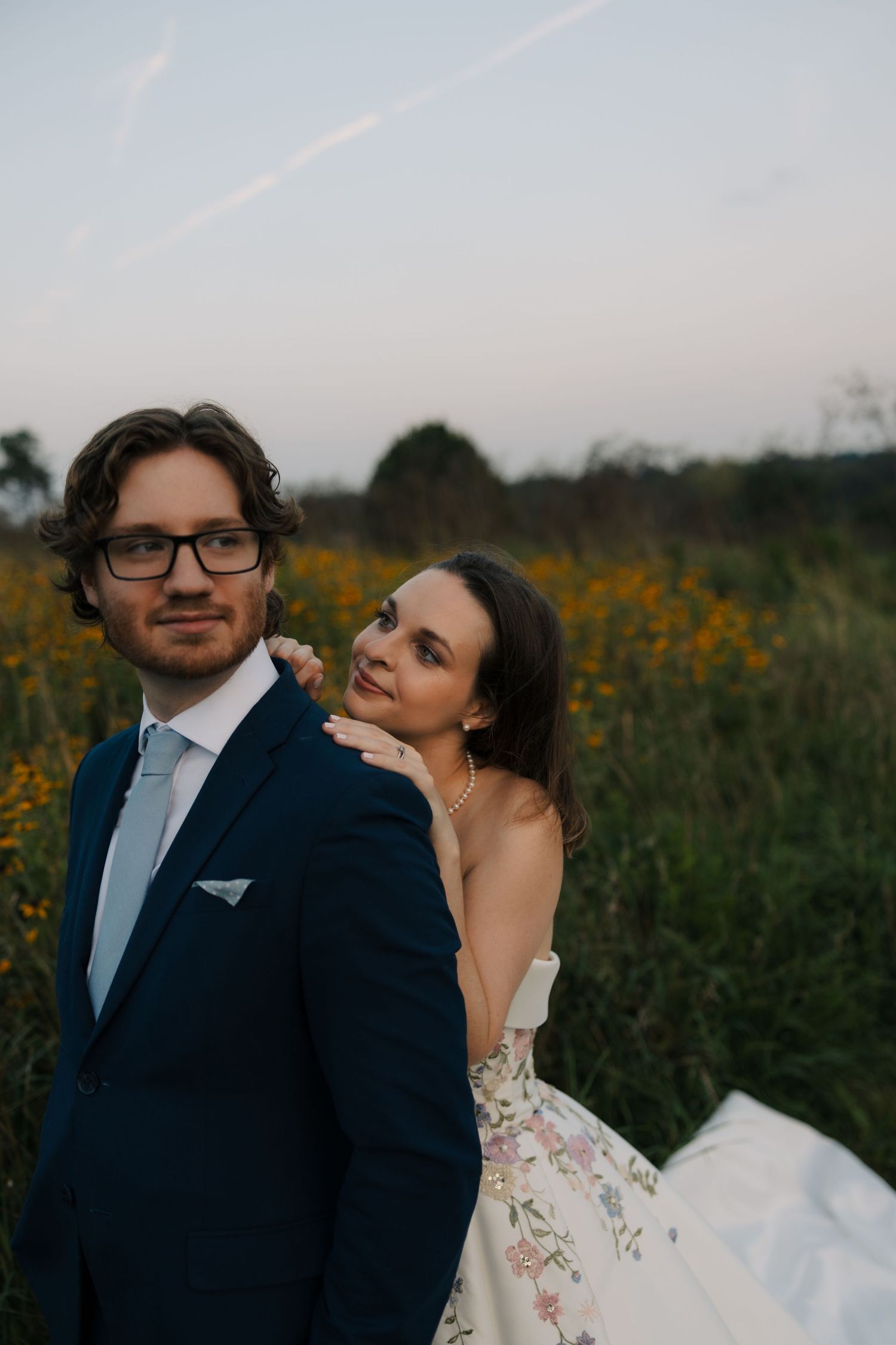 Groom in navy suit stands as bride in floral wedding gown leans on his shoulder in a wildflower field.