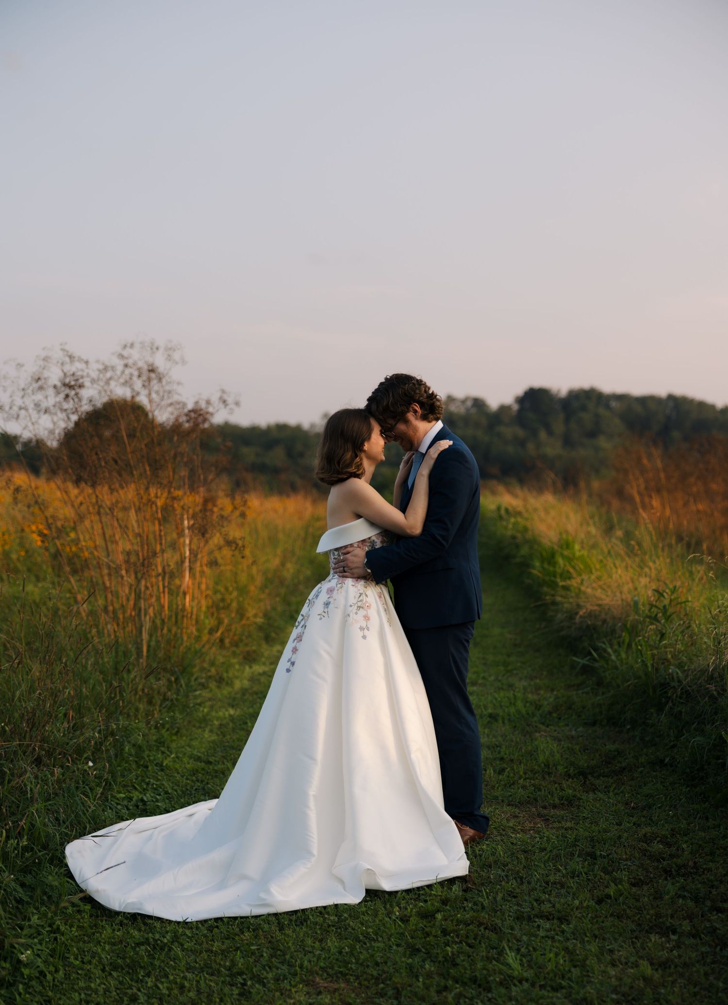 Bride and groom share a romantic forehead touch in a golden field at sunset during their outdoor wedding.