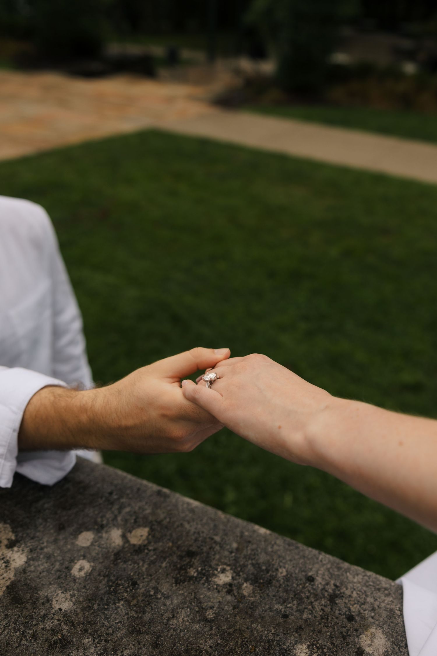 Close-up of couple holding hands revealing a delicate engagement ring against a lush green lawn.