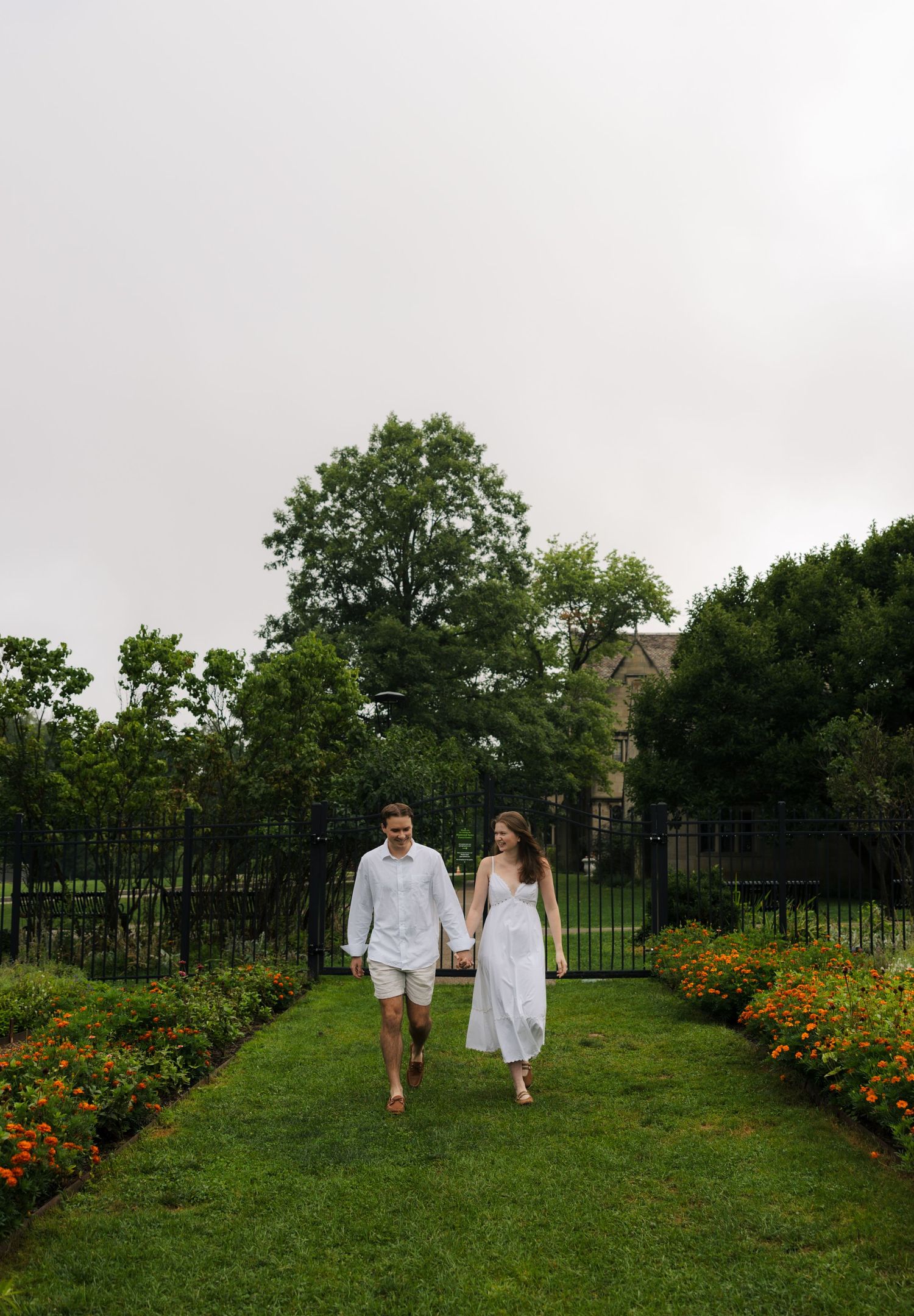 Couple in white outfits holding hands walking along a lush garden path lined with orange flowers and green trees.
