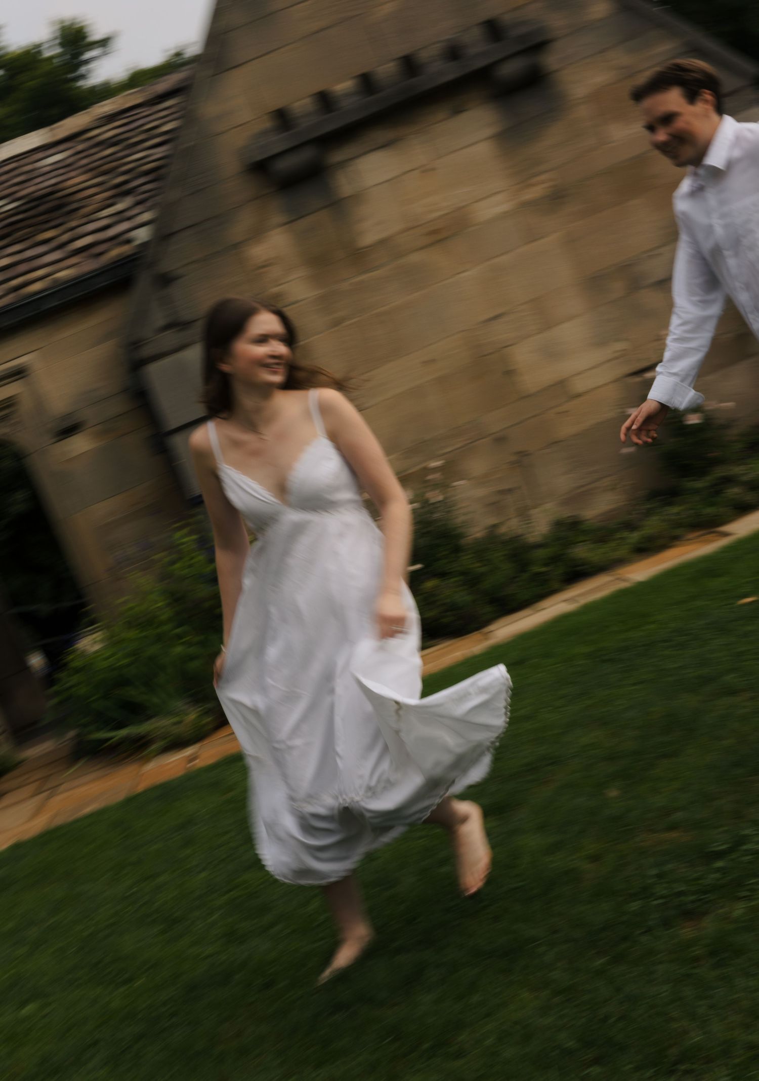 Motion-blur photo of joyful woman in white dress running on grass with man chasing her.