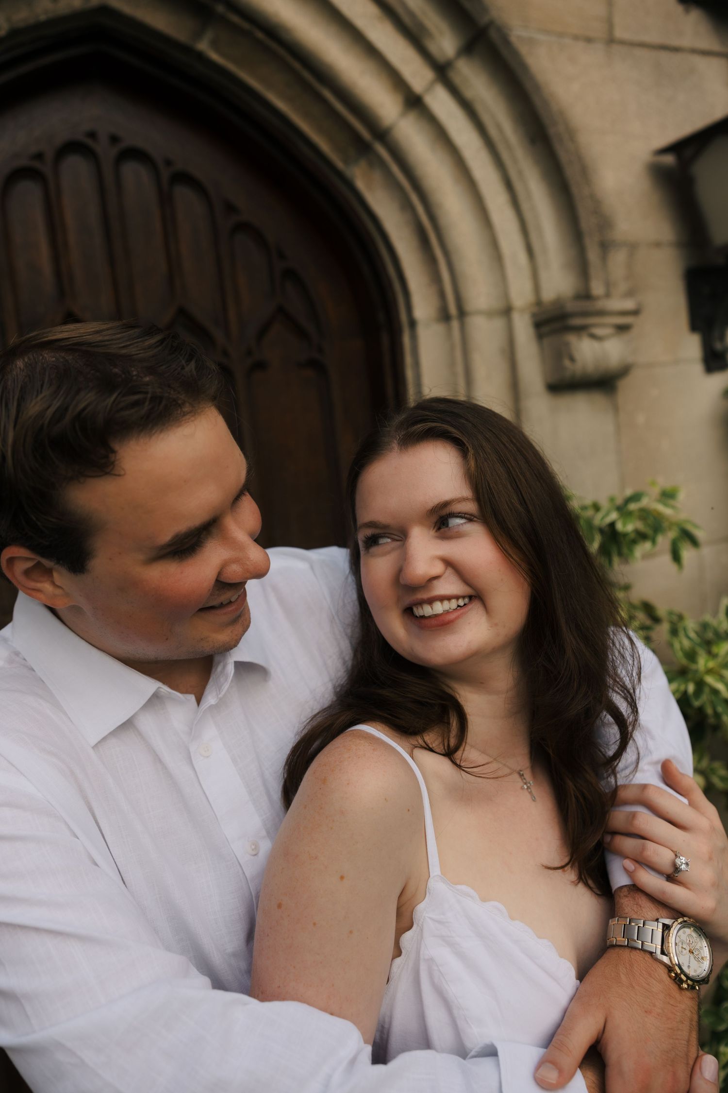 Happy couple sharing a tender glance beside gothic stone church doorway, woman wearing elegant watch.