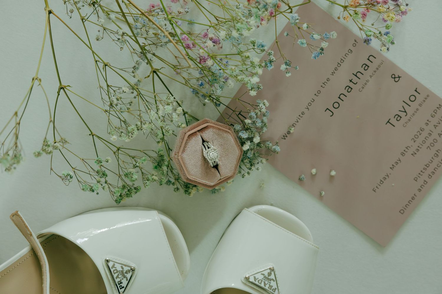 Wedding flat lay featuring a diamond ring in a wooden box, floral stems, white heels, and a Jonathan & Taylor invitation.