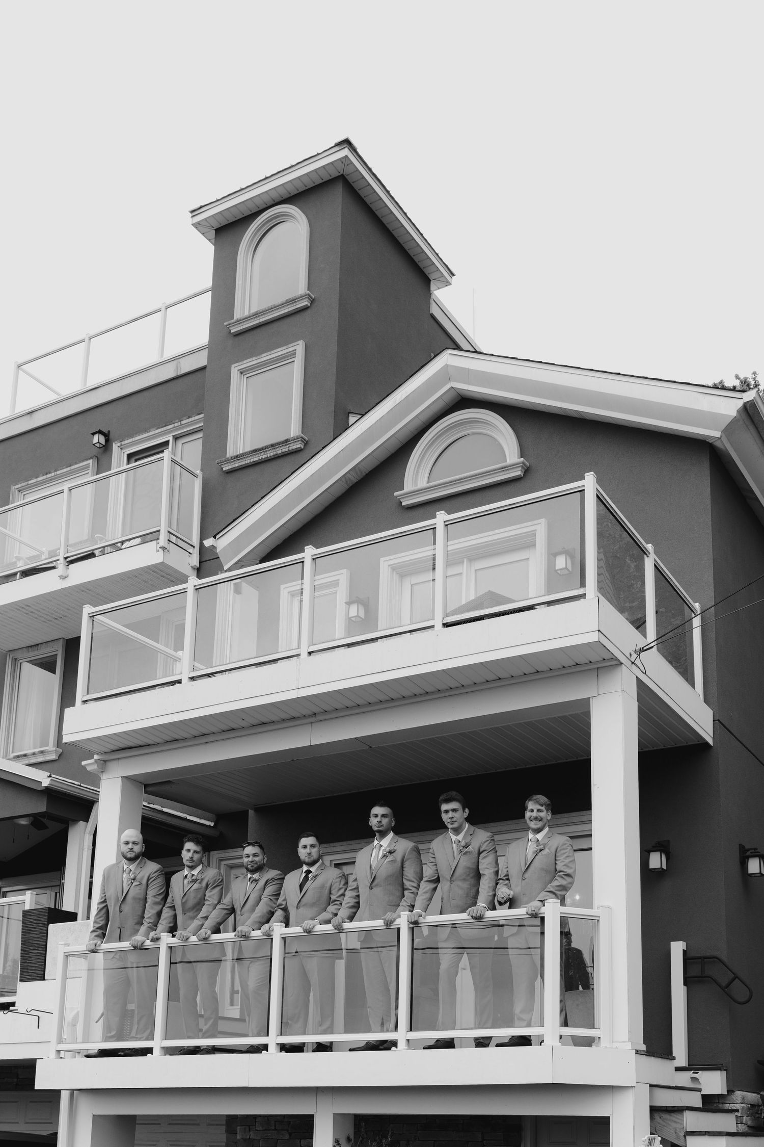 Groomsmen in suits standing on a balcony of a multi-story building in a black and white wedding photo.