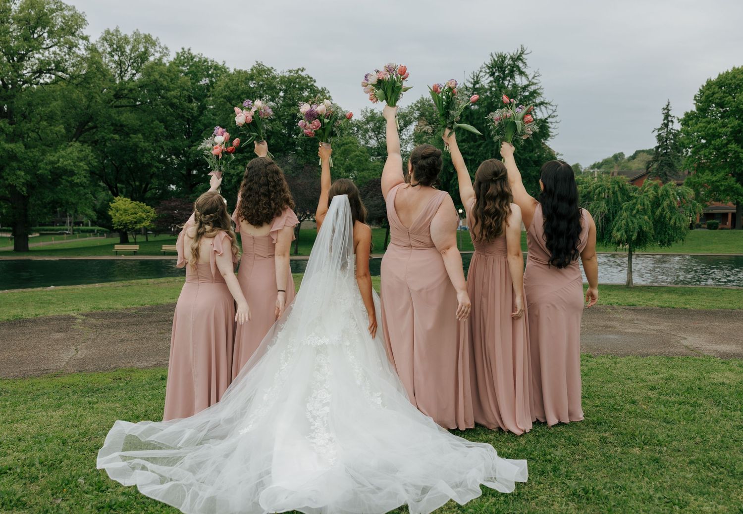 Bride and bridesmaids in blush dresses raise bouquets outdoors with lush greenery in the background.