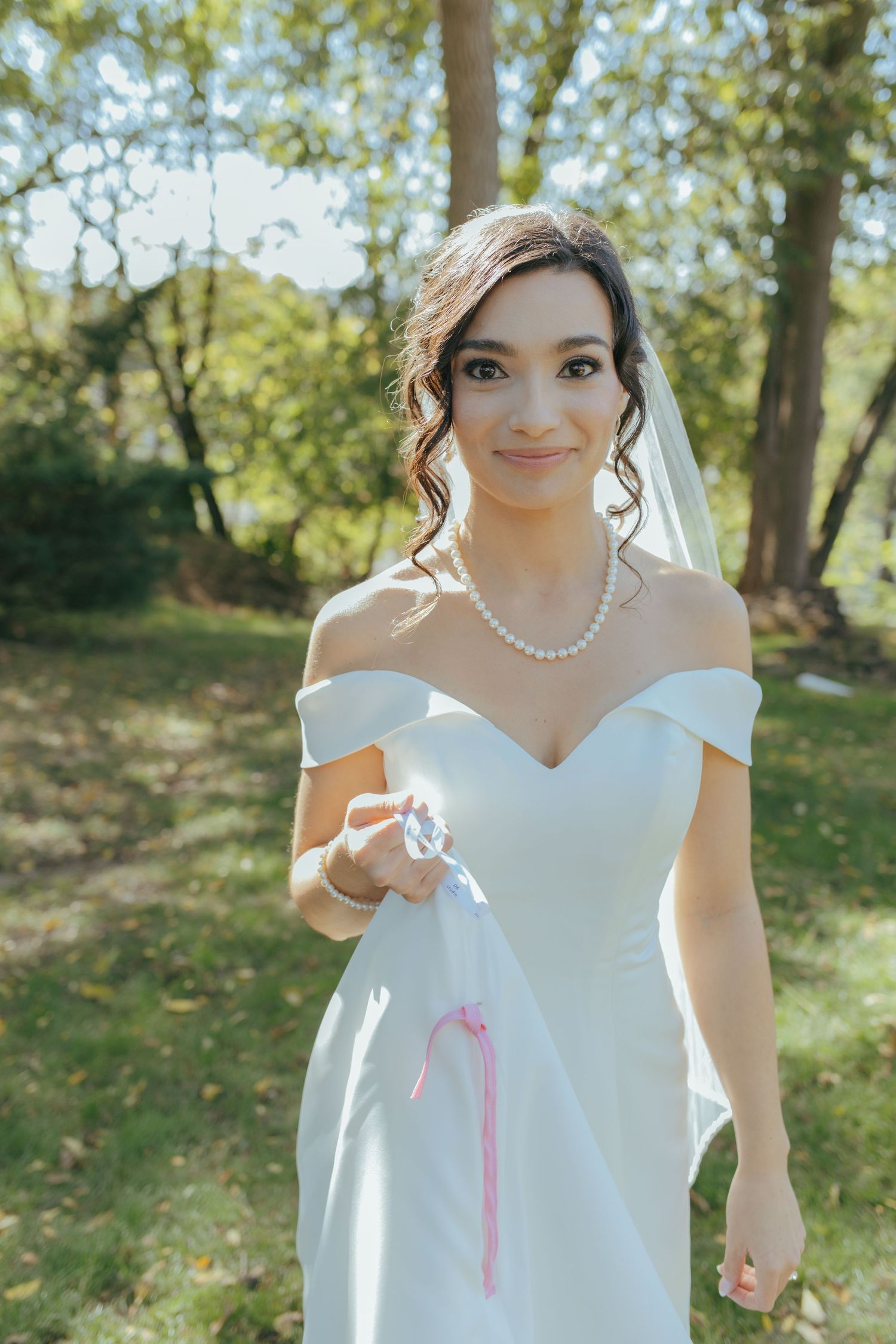 Elegant bride in off-shoulder white gown and pearl necklace holds bouquet in sunlit garden setting.