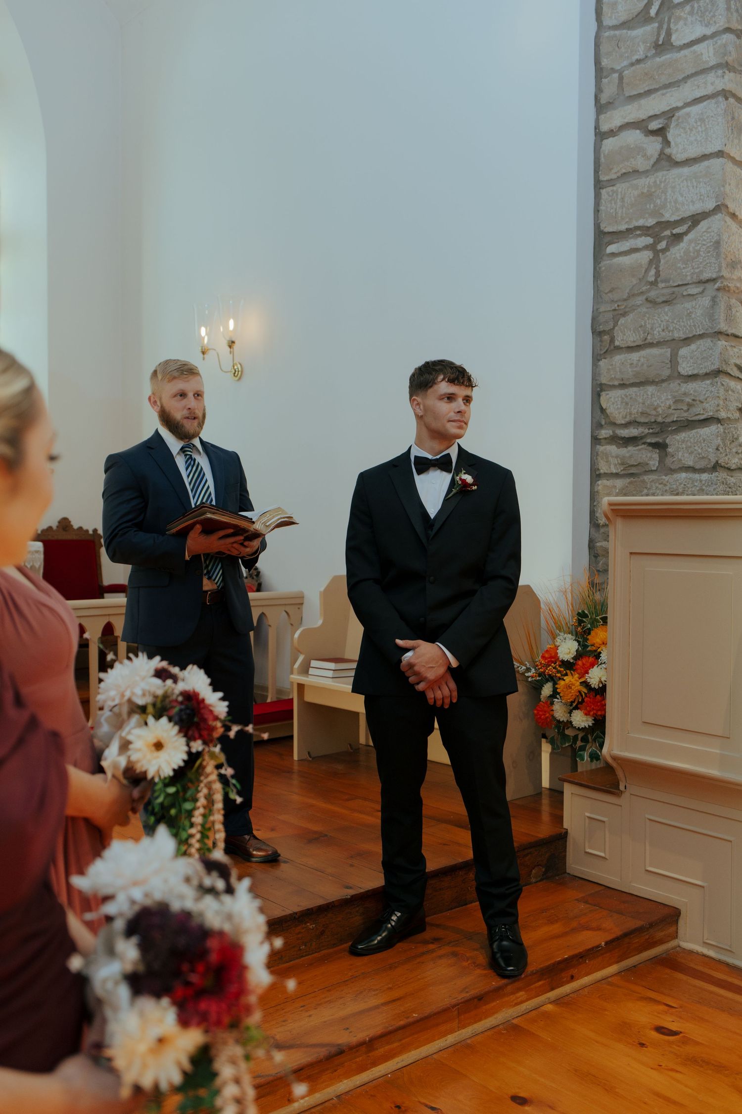 Groom in black tuxedo stands at wedding altar as officiant holds book and bridesmaid holds bouquet.