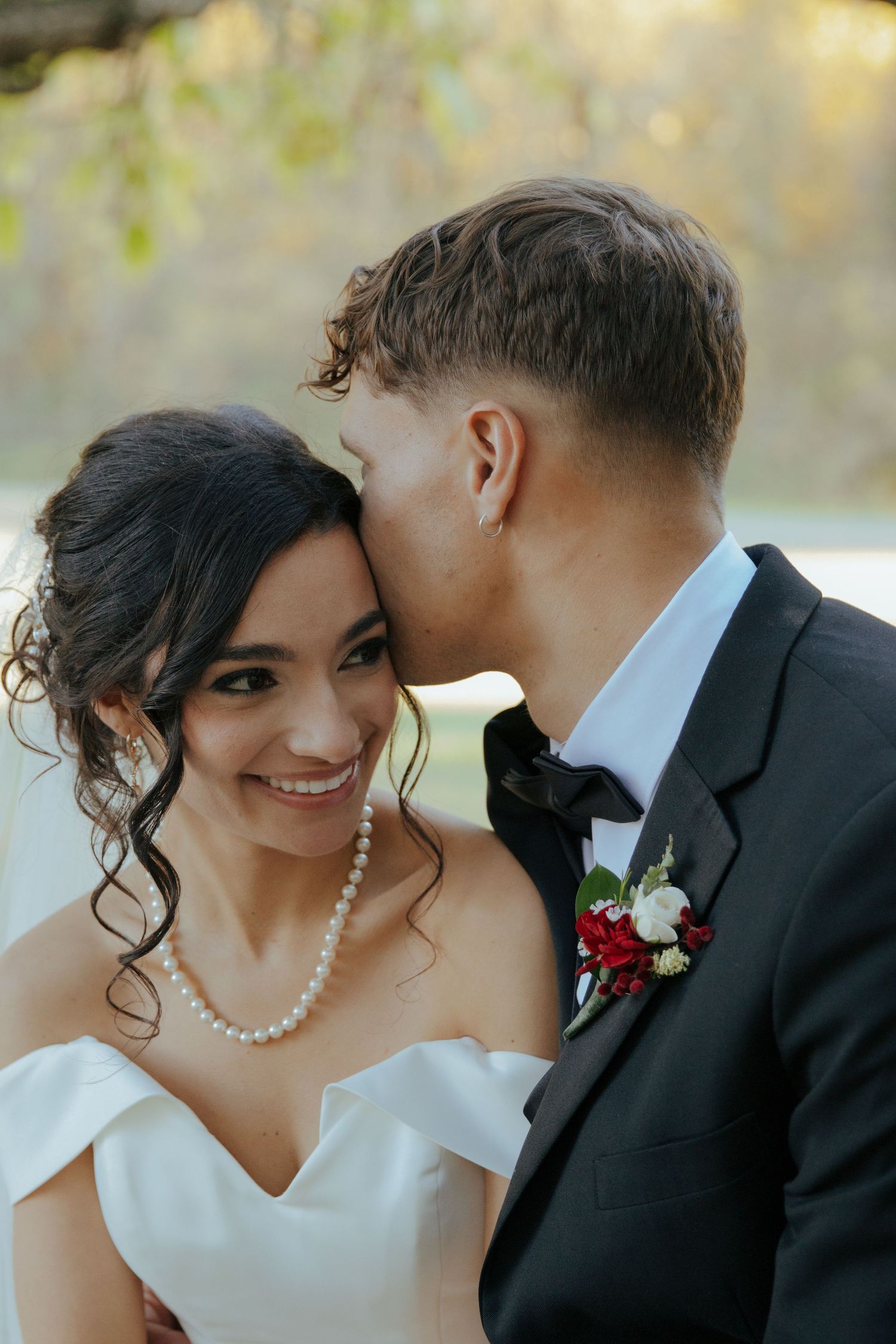 Groom in black tuxedo tenderly kissing smiling bride wearing pearl necklace and veil in soft outdoor autumn light.