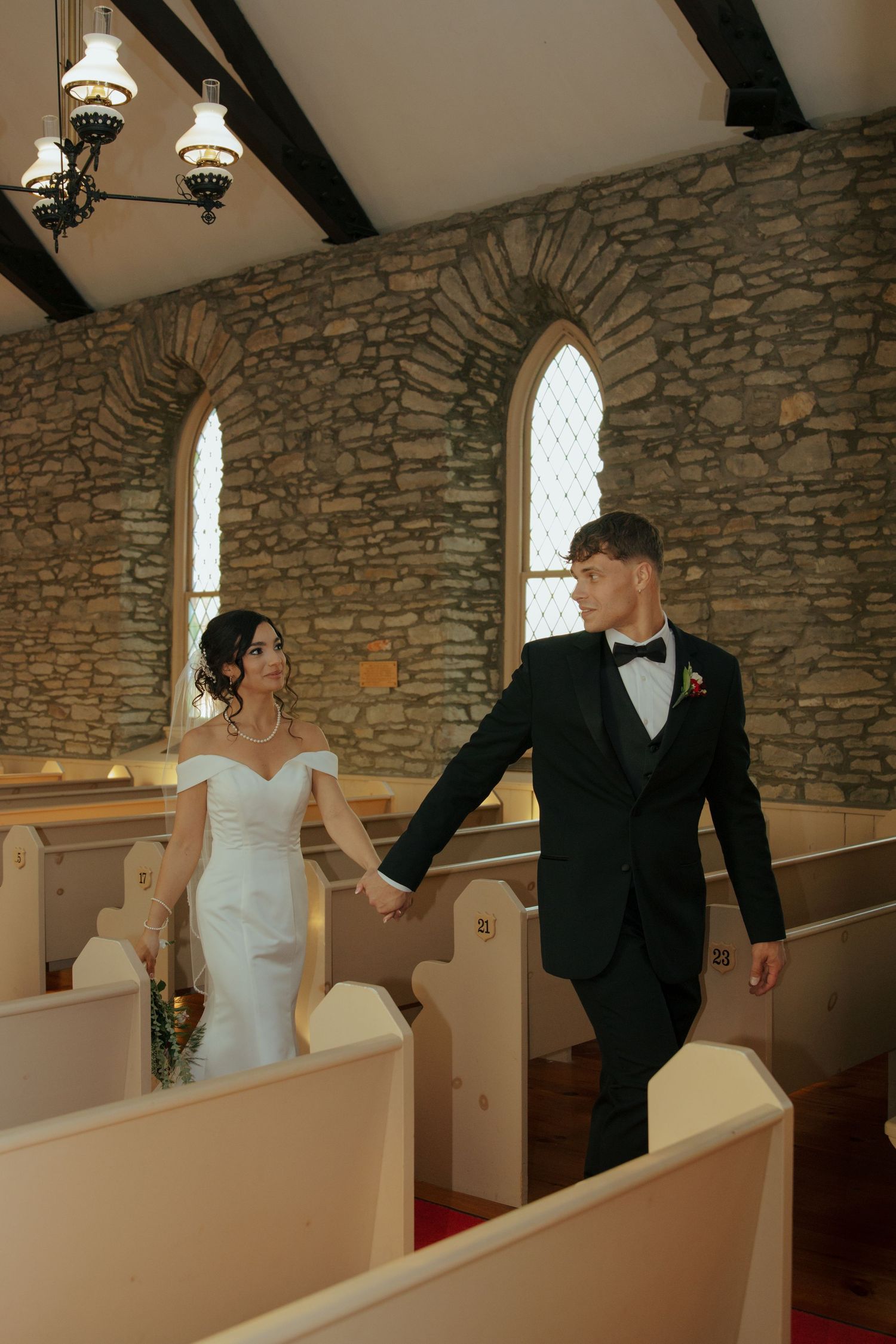 Bride and groom walk hand in hand down aisle of rustic stone chapel with wooden pews.