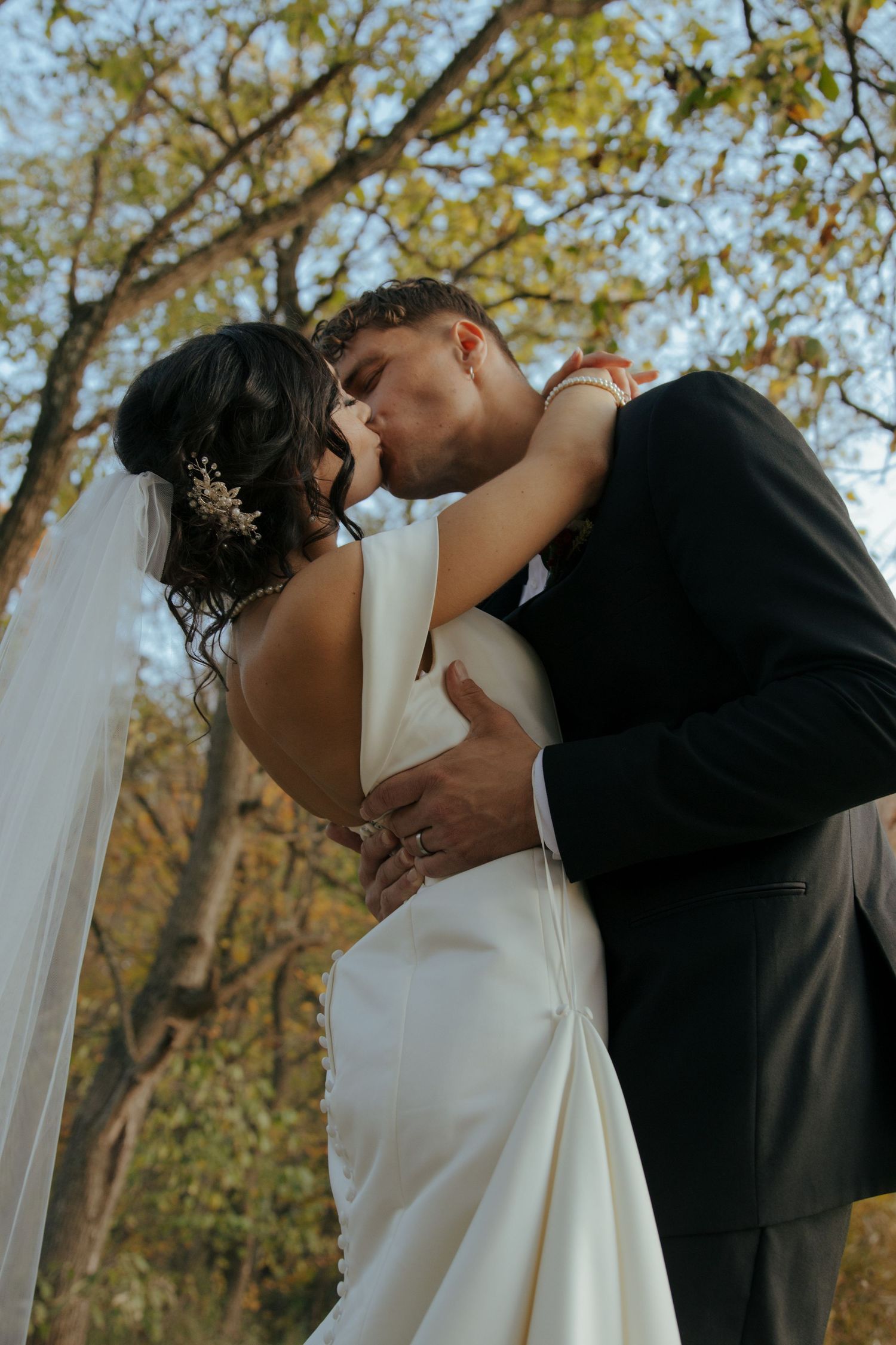 Bride and groom share romantic first kiss outdoors surrounded by autumn trees in elegant wedding attire.
