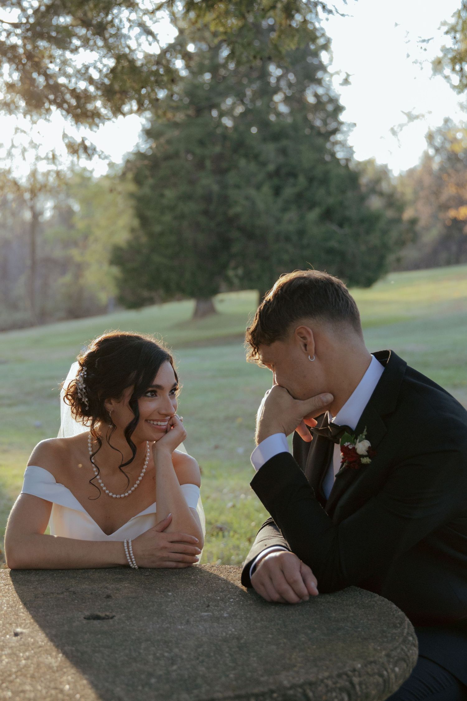 Bride and groom share an intimate moment leaning on a stone surface in a sunlit outdoor park setting.