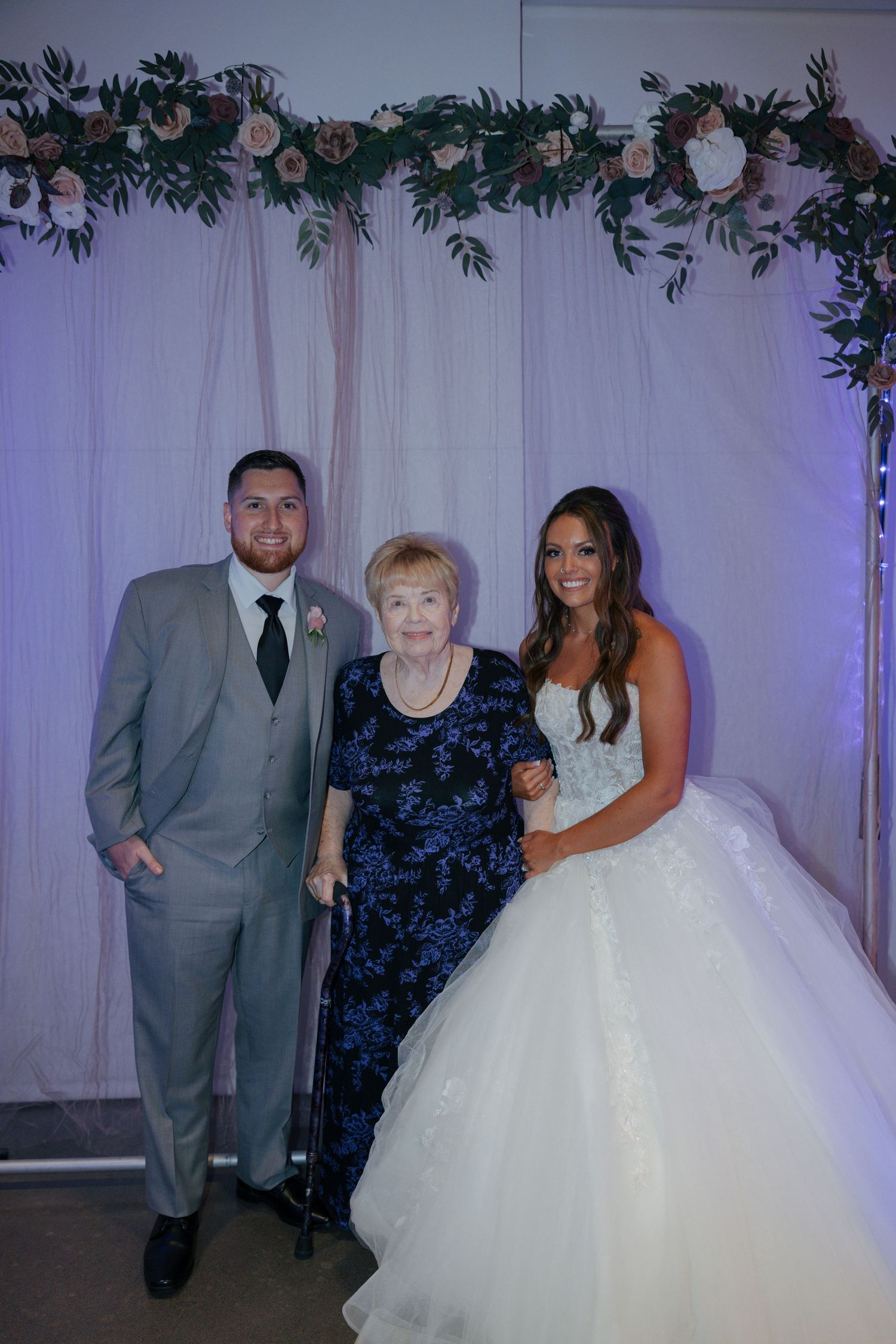 Bride and groom pose with elderly grandmother in front of floral backdrop at wedding reception.