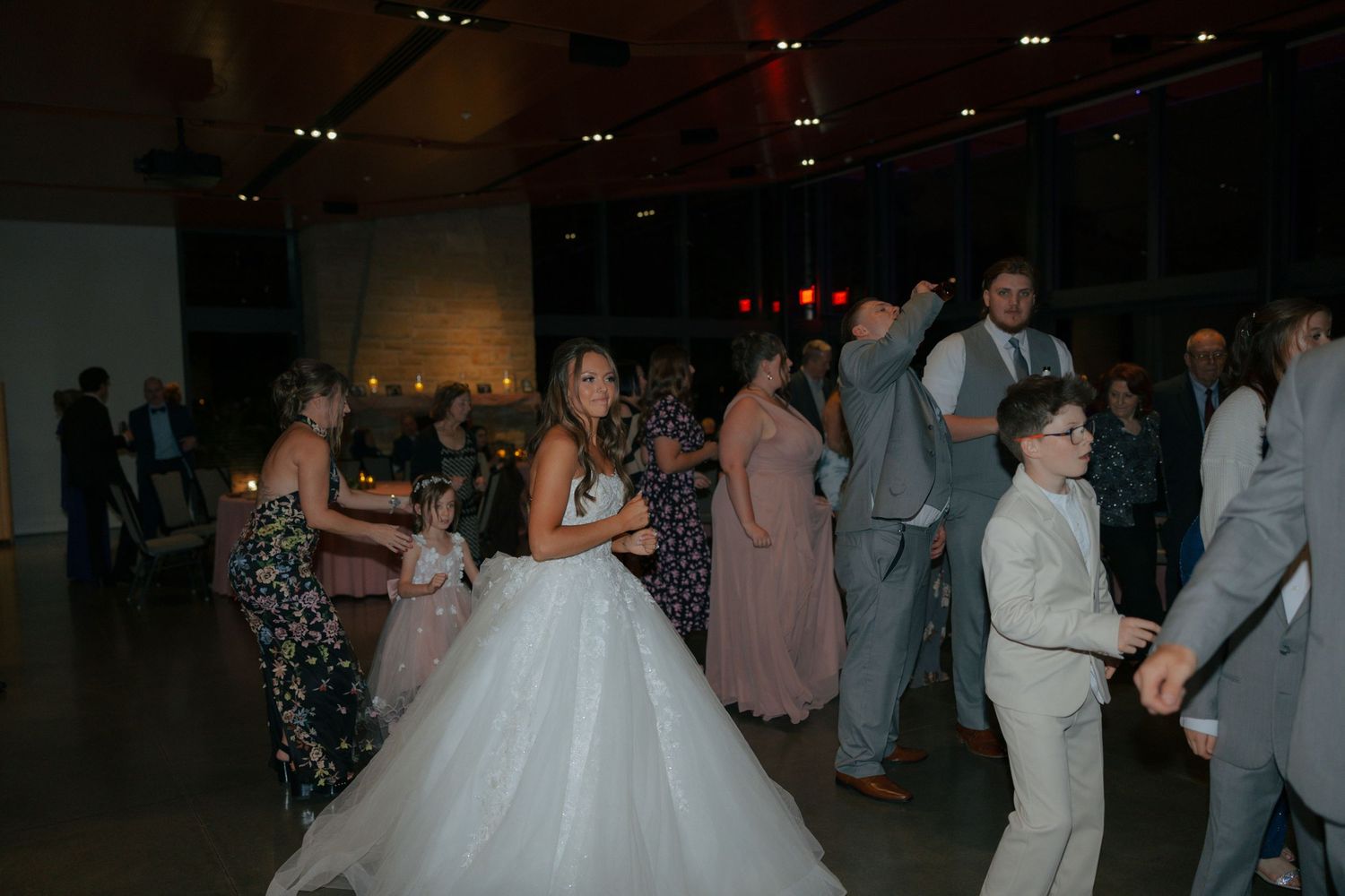Bride in white ball gown dancing with guests including a young boy in gray suit at a lively wedding reception.