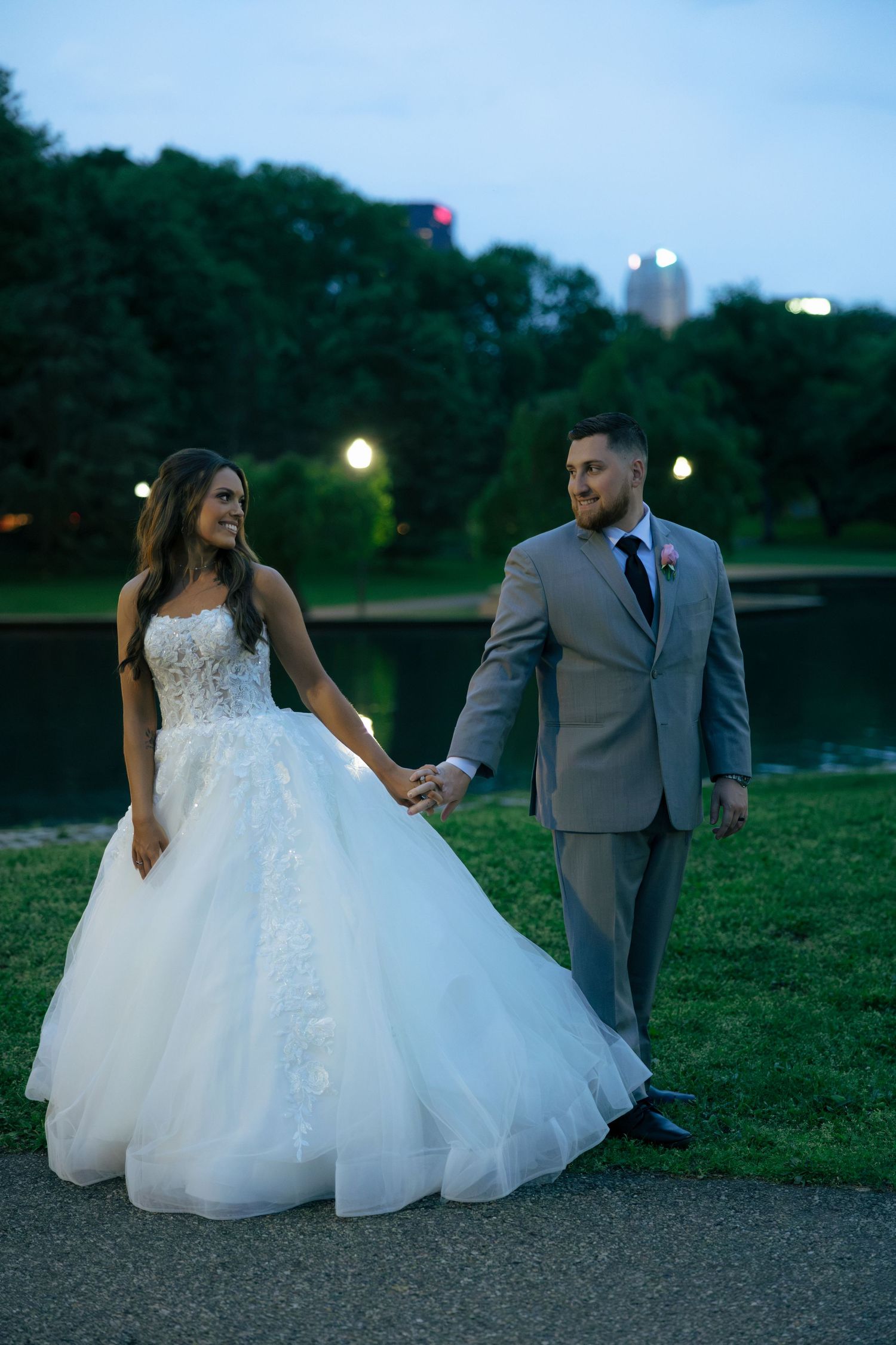 Newlyweds walk hand-in-hand in a park at dusk, bride in a flowing white ballgown.