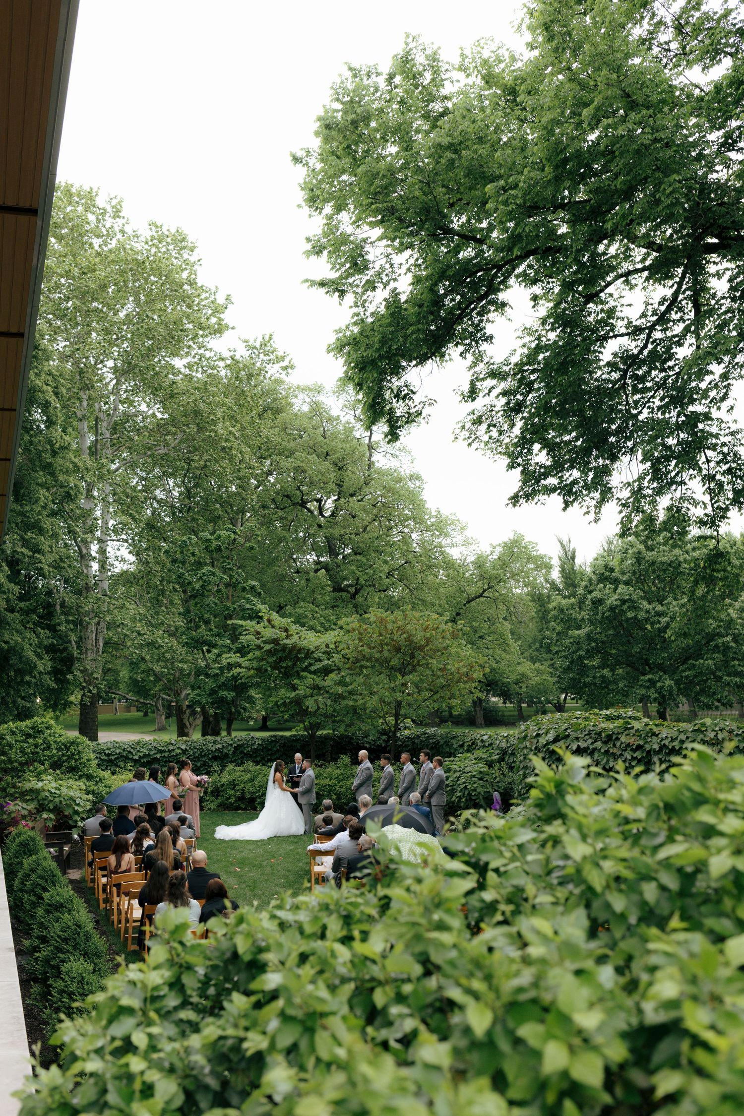 Aerial view of outdoor wedding ceremony surrounded by lush green trees and manicured hedges.
