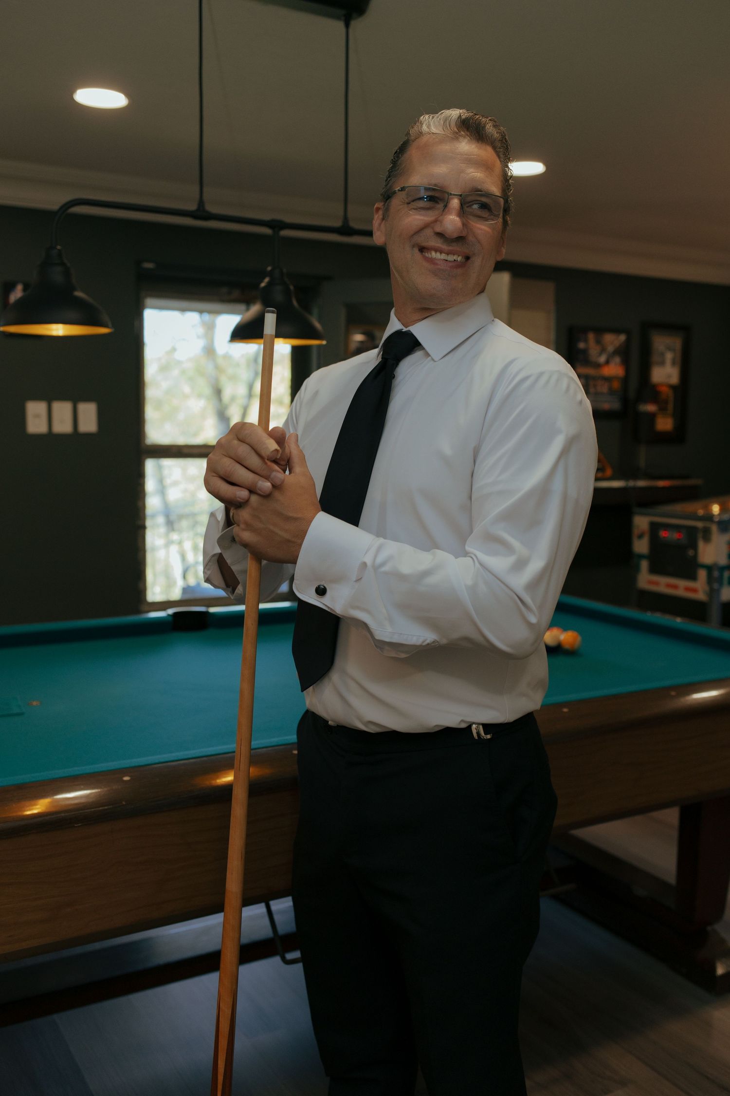 Smiling man in white shirt and tie posing with pool cue beside billiard table at wedding.