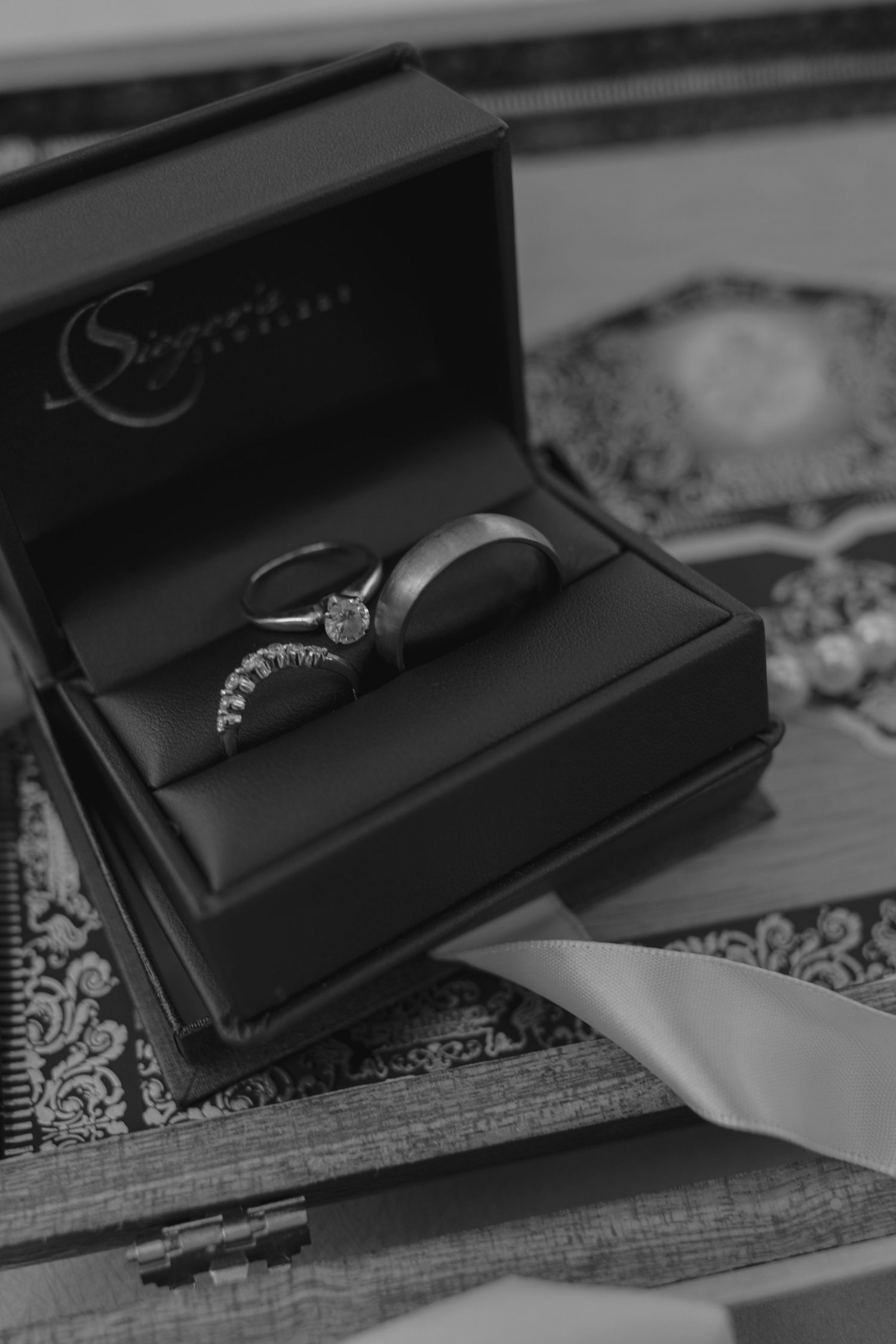 Black and white photo of wedding rings in a jewelry box, featuring a diamond engagement ring and a plain band.