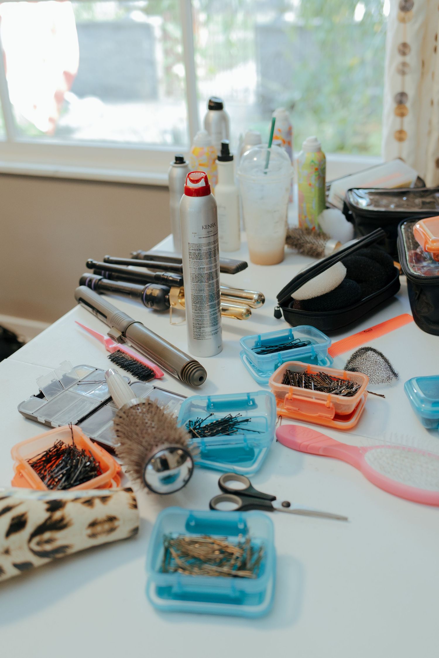 Hair styling tools, bobby pins, scissors, and hair spray spread across a white table for wedding preparation.