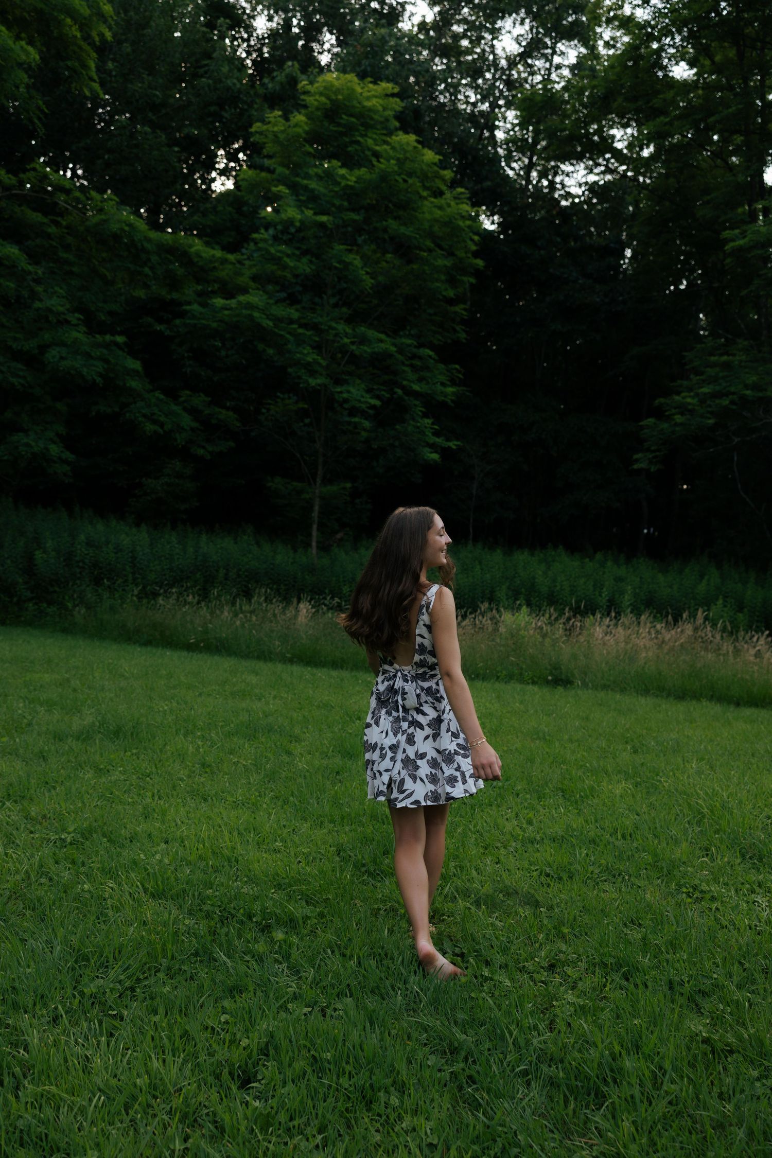 Barefoot young woman in floral sundress standing on green grass in wooded park.