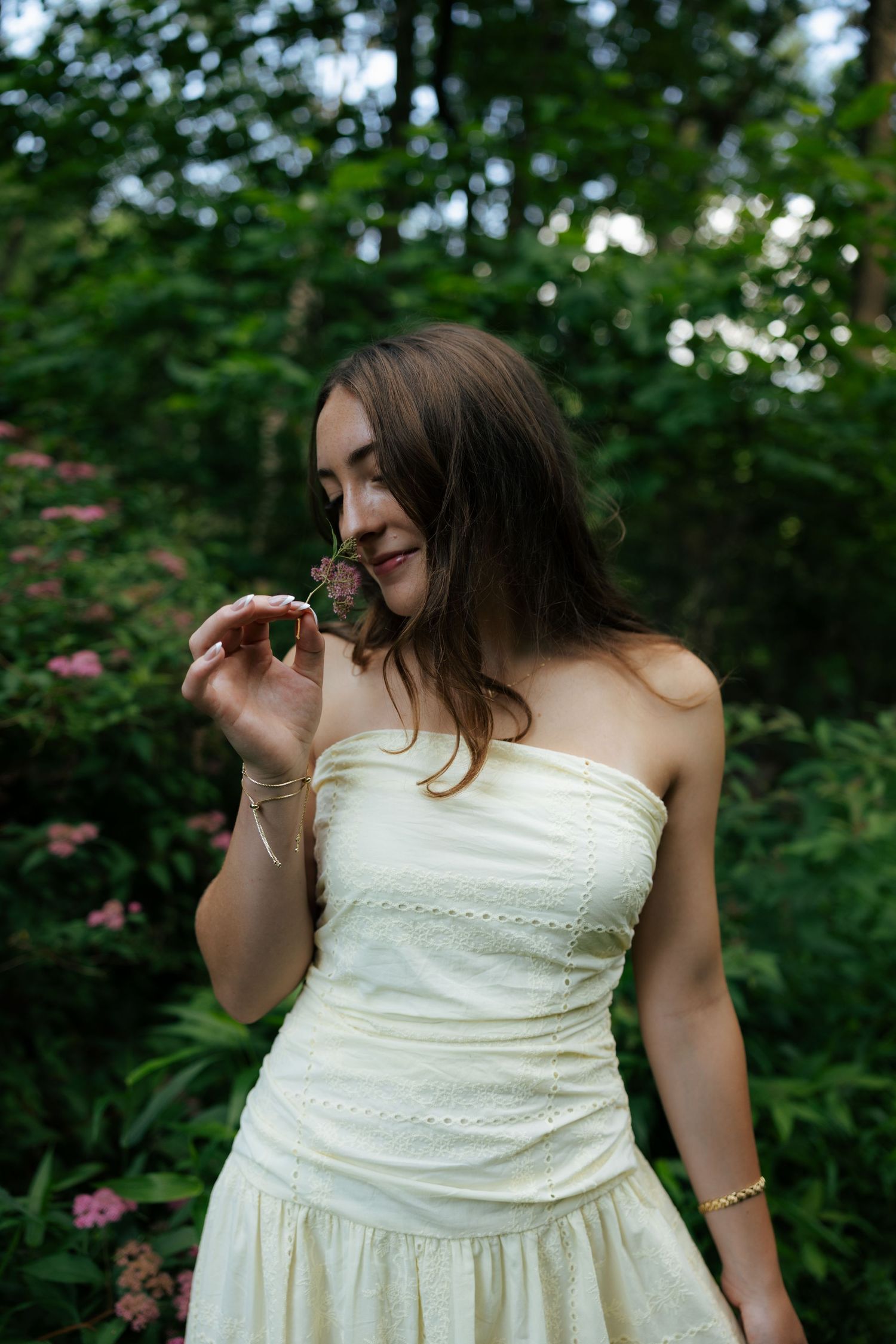 Young woman in white strapless dress smelling a flower in a lush green garden with pink blooms.
