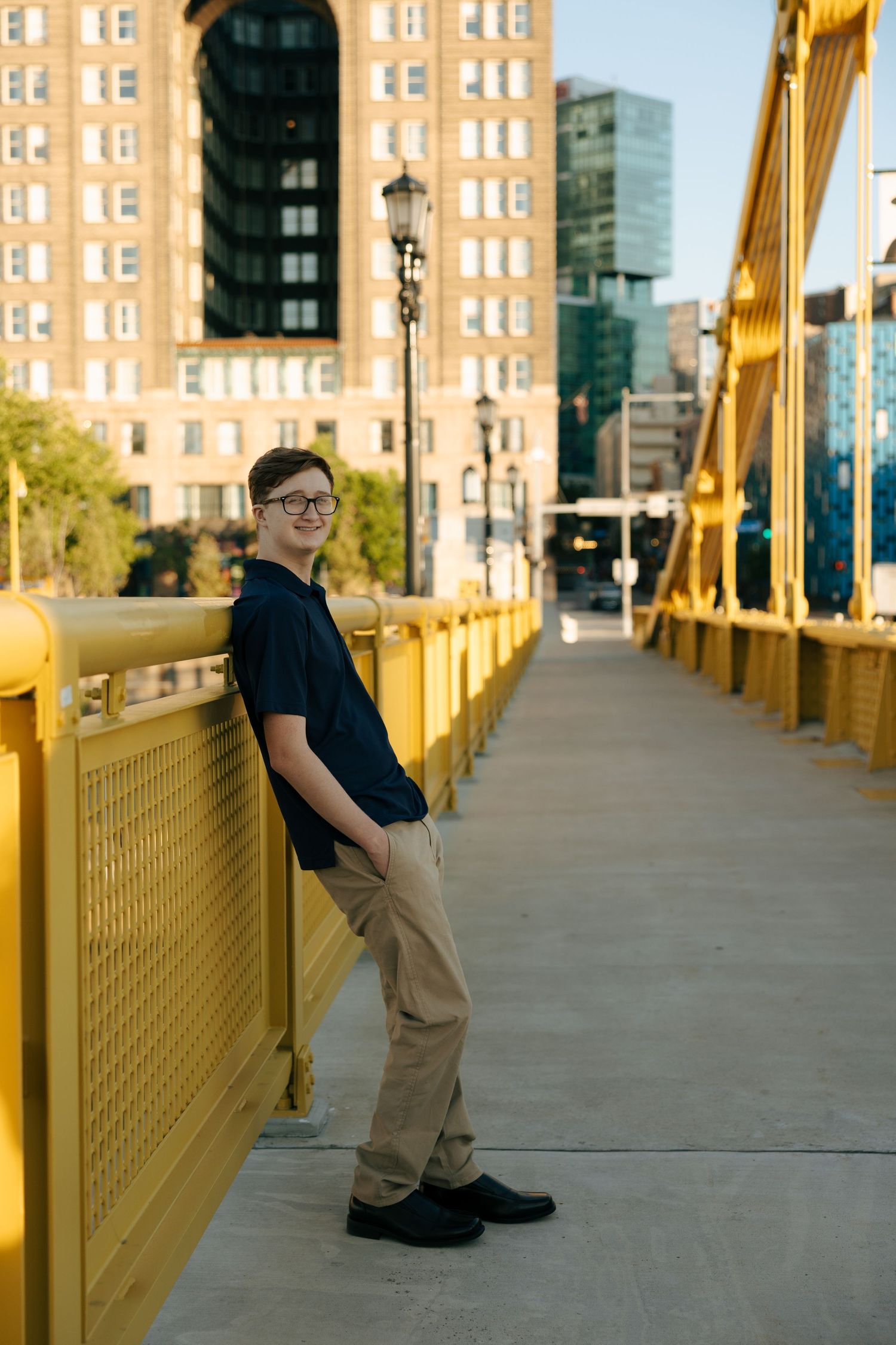 Teen boy in glasses leans on yellow bridge railing in urban Pittsburgh setting, wearing navy shirt and khaki pants.
