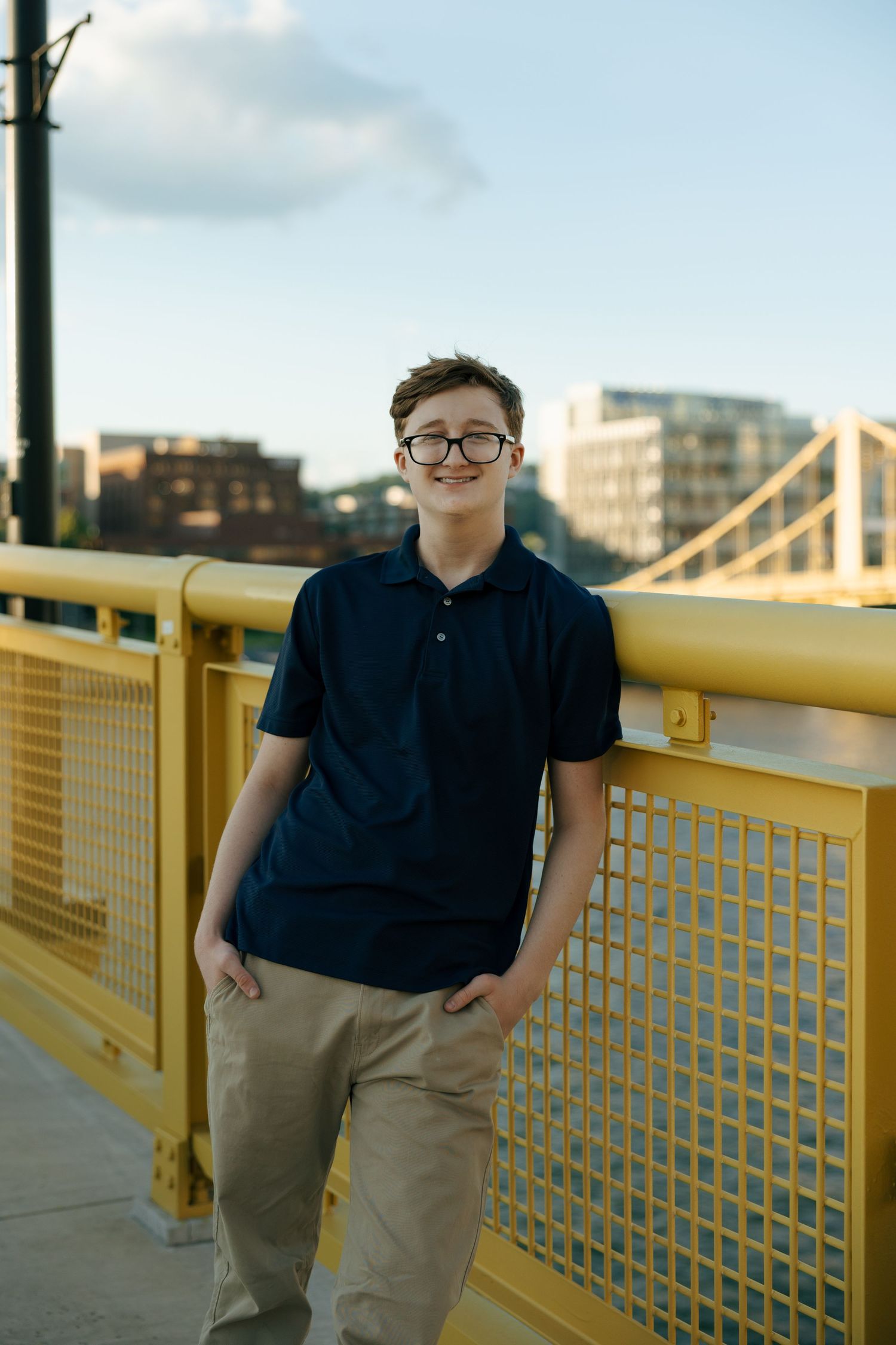 Young man in navy polo and khakis posing on yellow bridge with hands in pockets and city view.