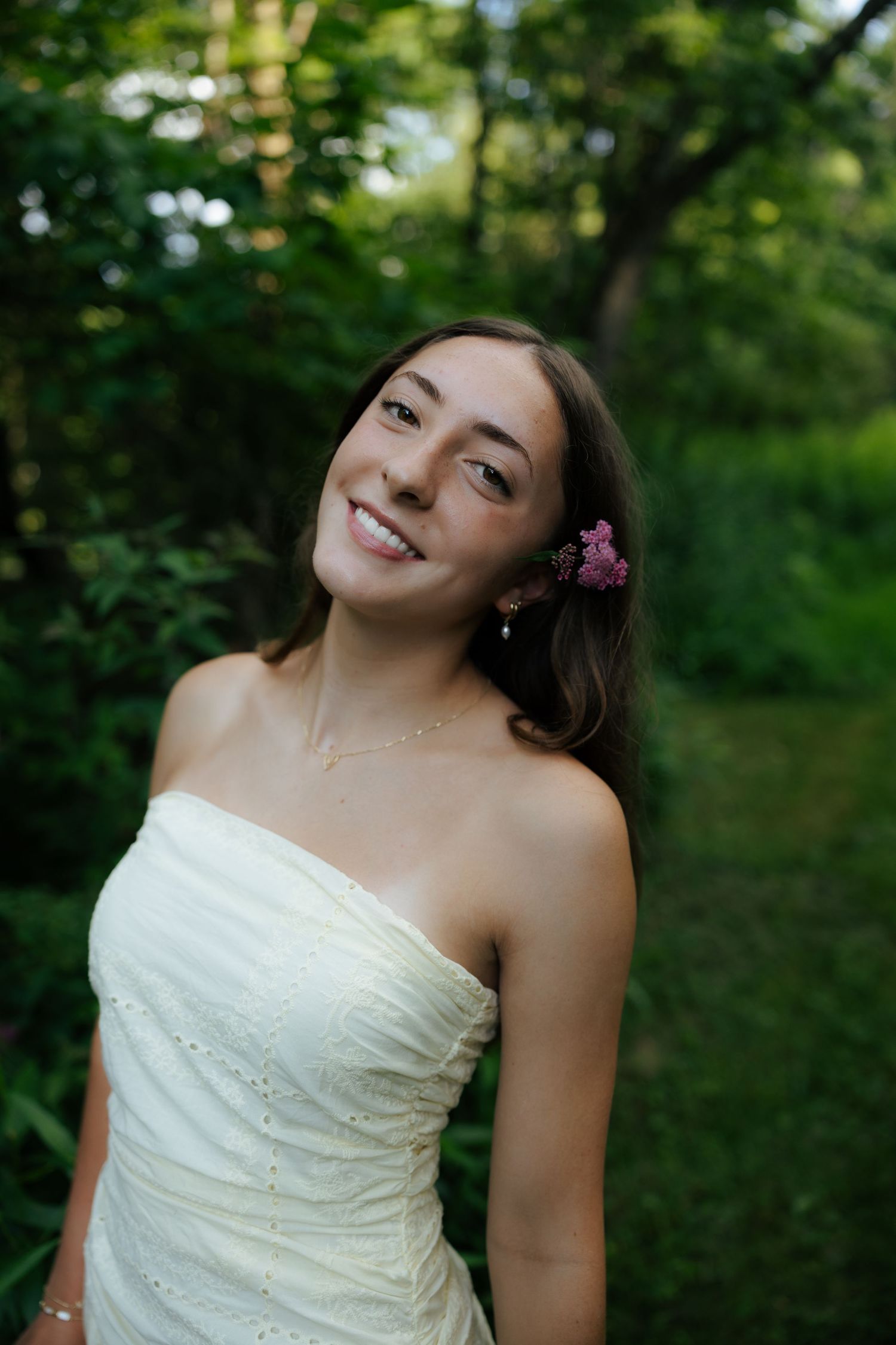 Smiling brunette woman in white strapless dress with pink flower in hair outdoors.