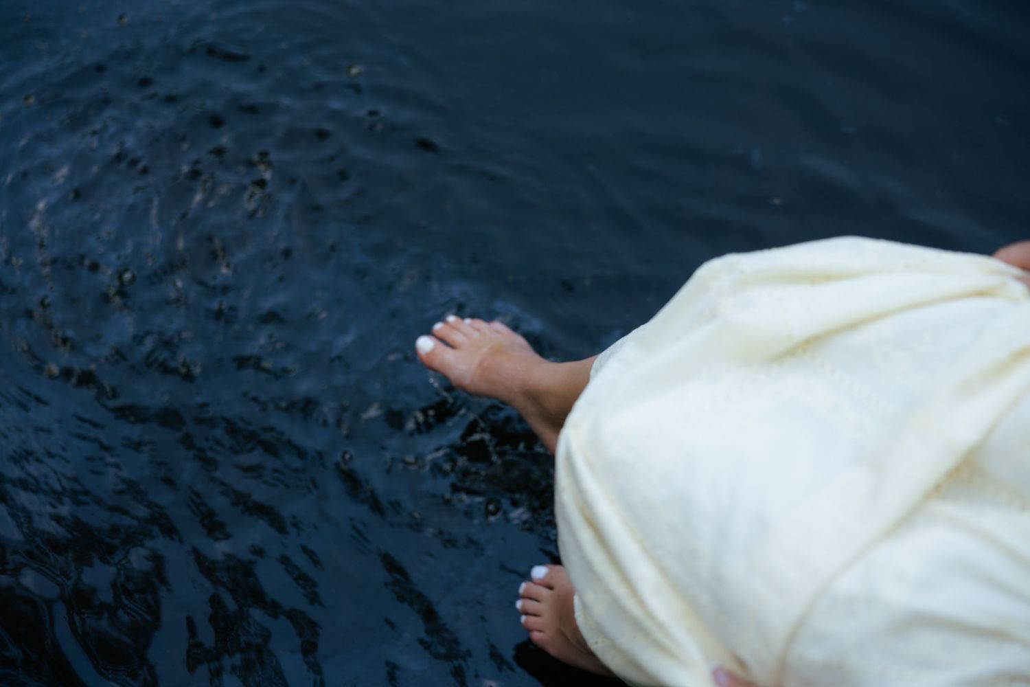 A person in a white dress dangles their bare feet over dark water from a wooden dock.