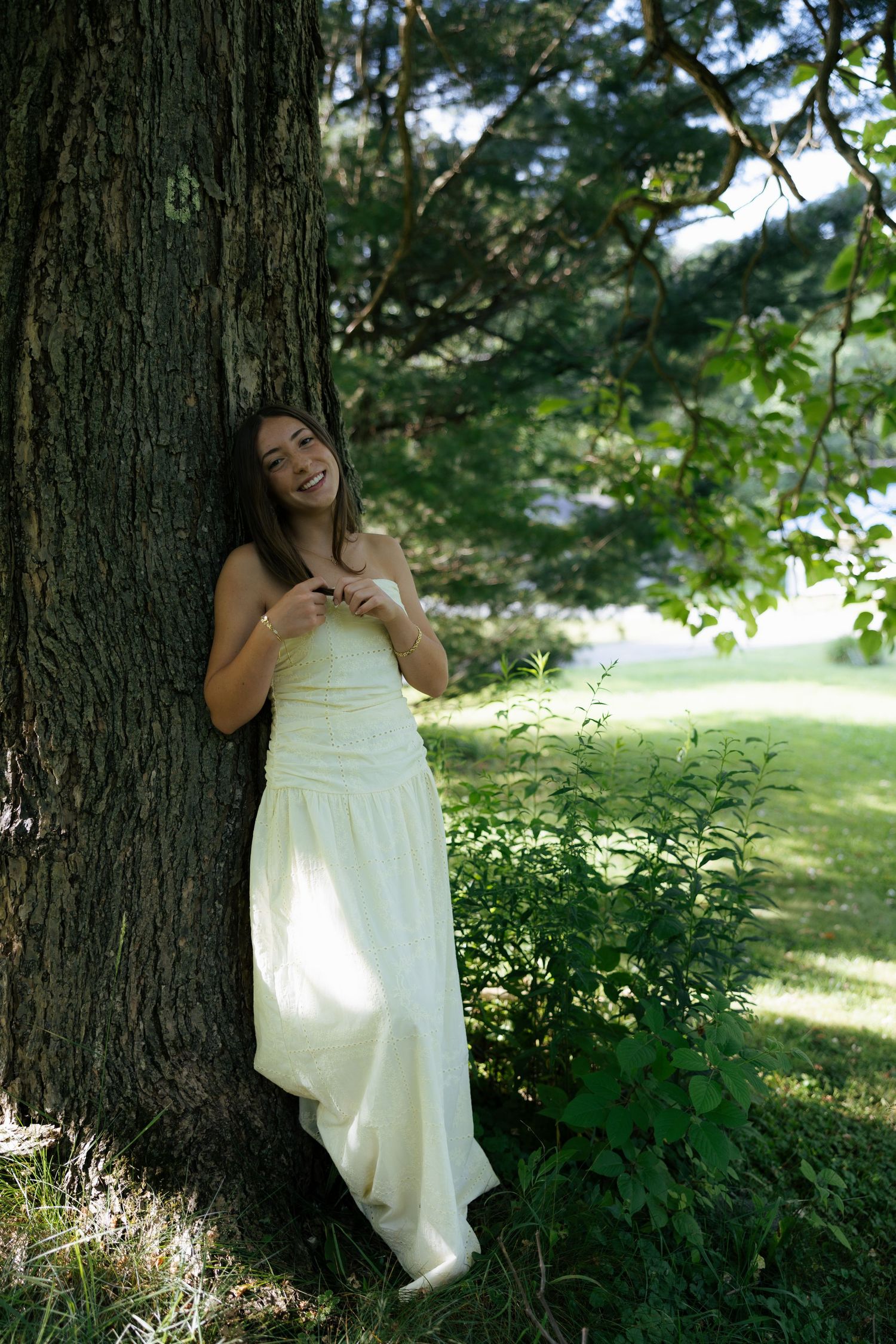 Young woman in white strapless gown leaning against large tree trunk smiling in lush green park.