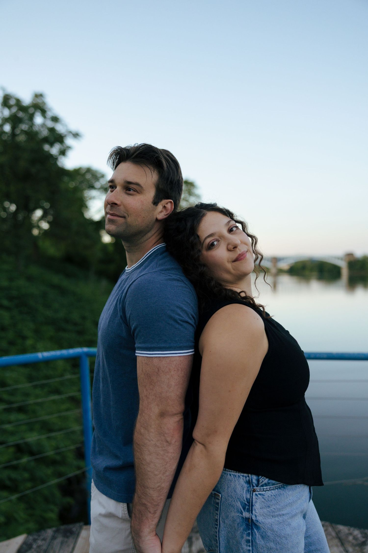 Couple standing back to back smiling by a river at dusk on a wooden bridge.