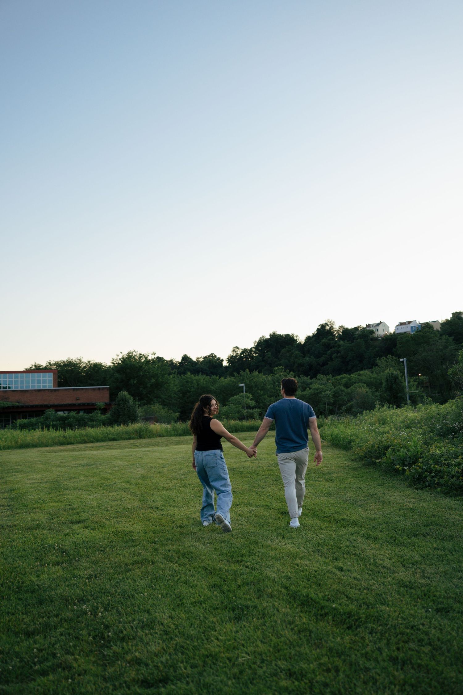 A couple holds hands while walking across a green lawn at dusk with trees and a hillside in the background.
