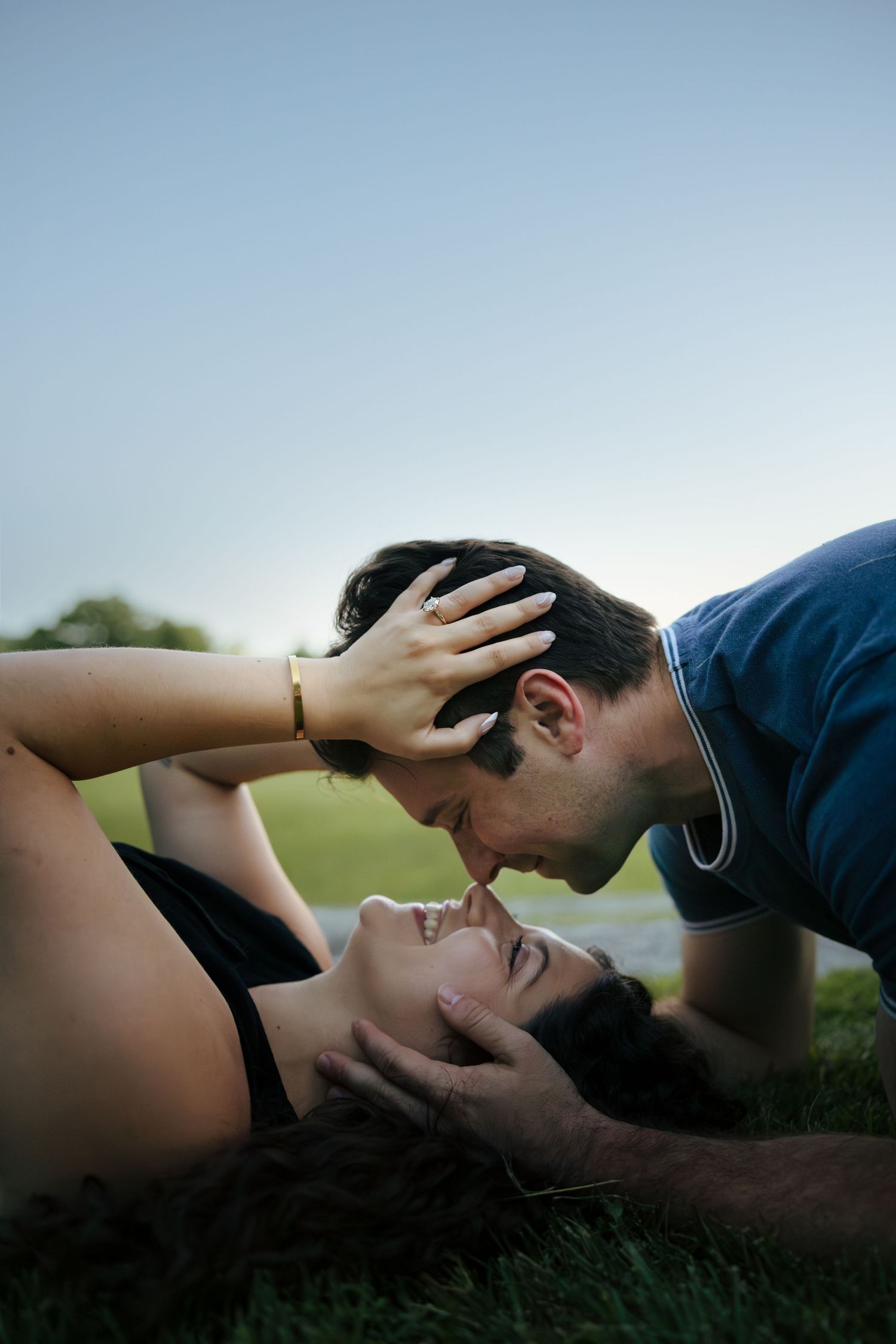 Romantic couple lying on grass about to kiss, woman wearing engagement ring at dusk outdoors.
