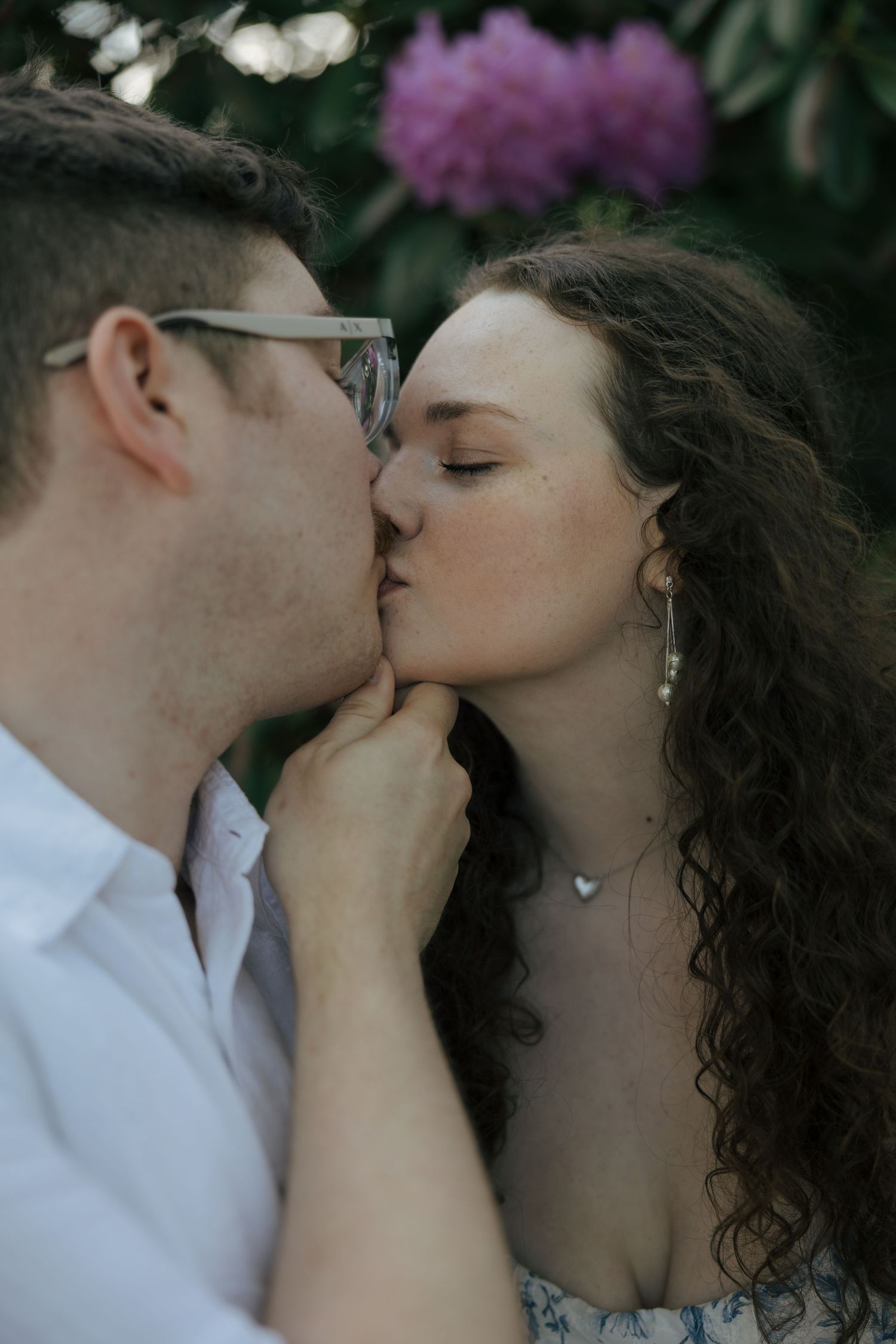 Engaged couple sharing a kiss outdoors with purple flowering bushes in the background during engagement session.