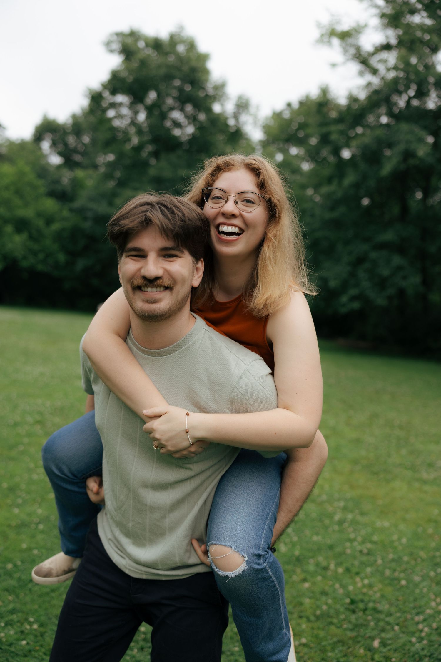 Laughing woman riding piggyback on smiling man with mustache in a lush green park.