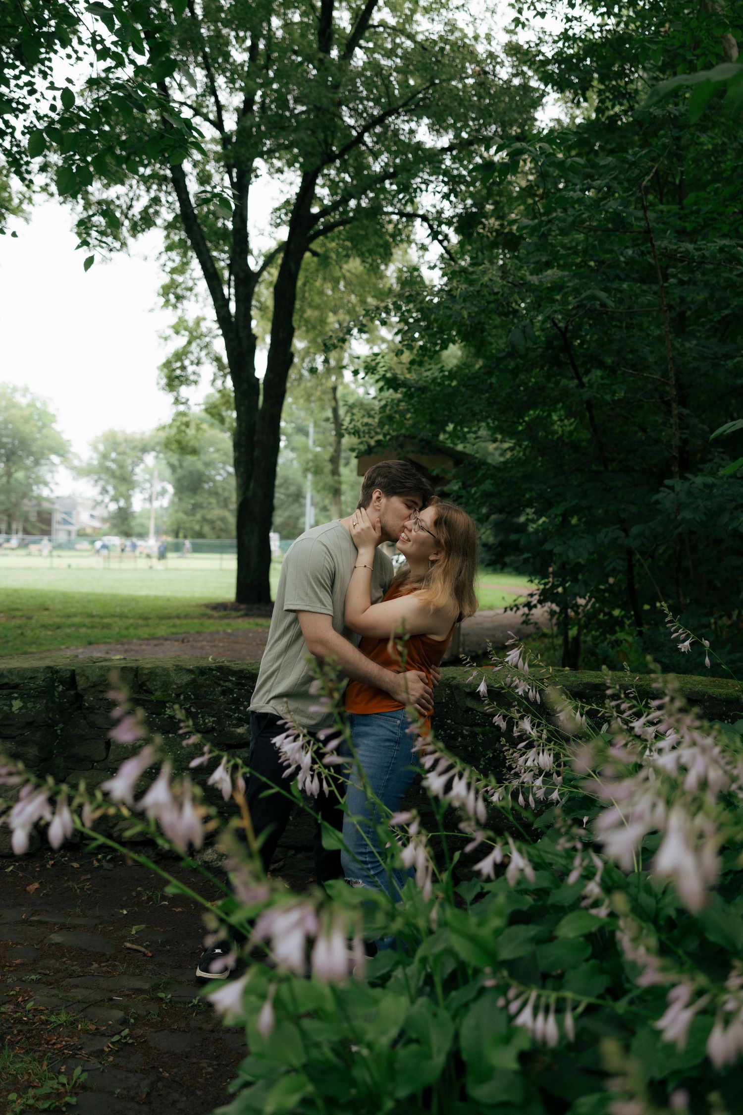 Couple sharing a kiss in a lush green park framed by blooming flowers in the foreground.
