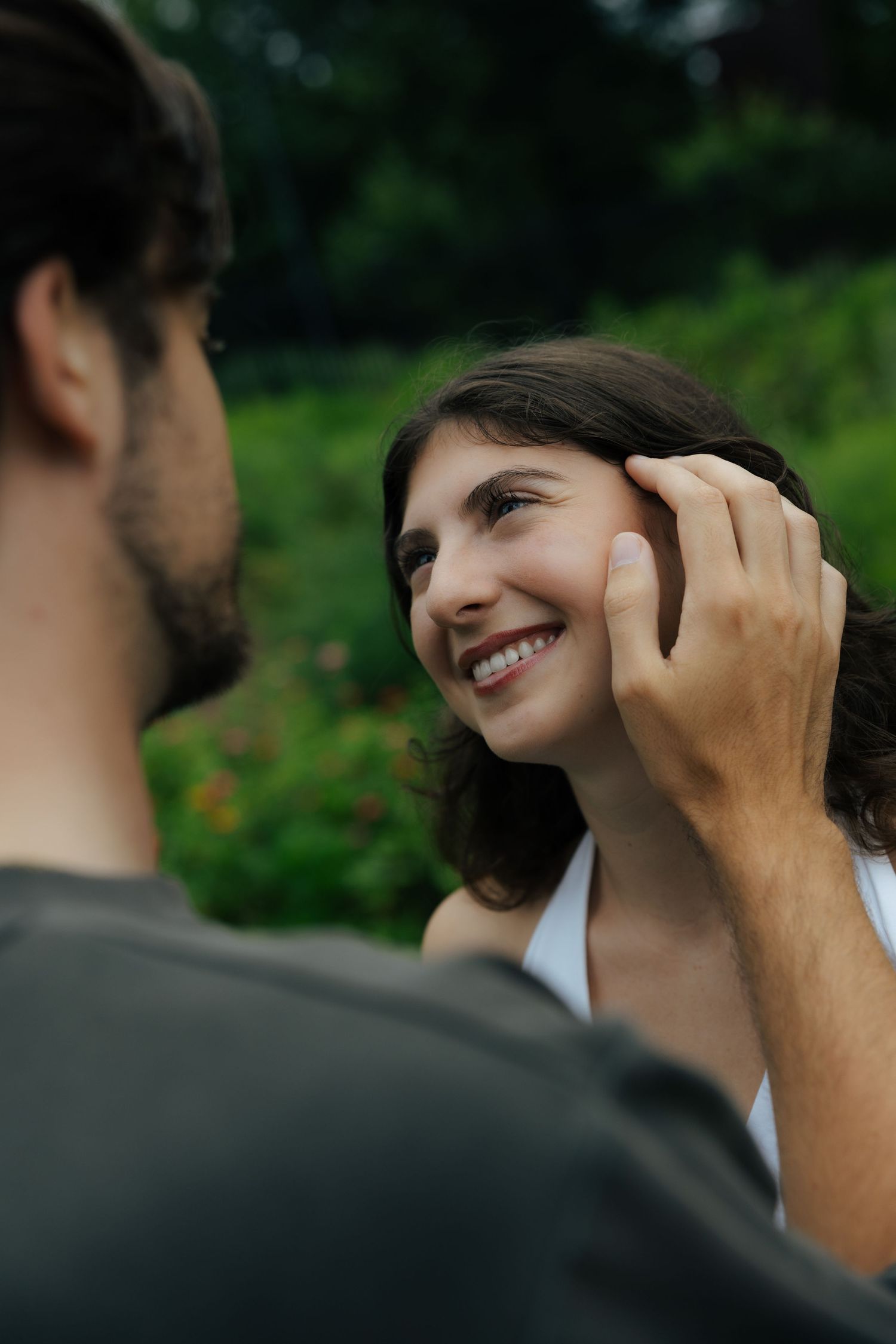 Young woman smiling as man gently touches her face outdoors in a lush green garden setting.