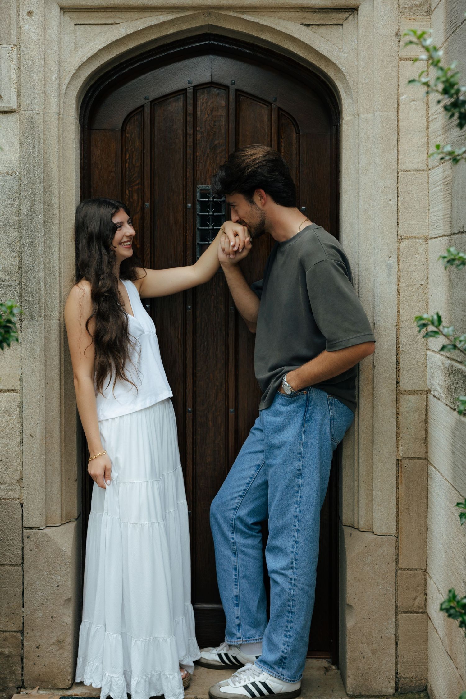 Man kissing woman's hand in front of ornate wooden arched door of stone building, woman in white dress smiling.