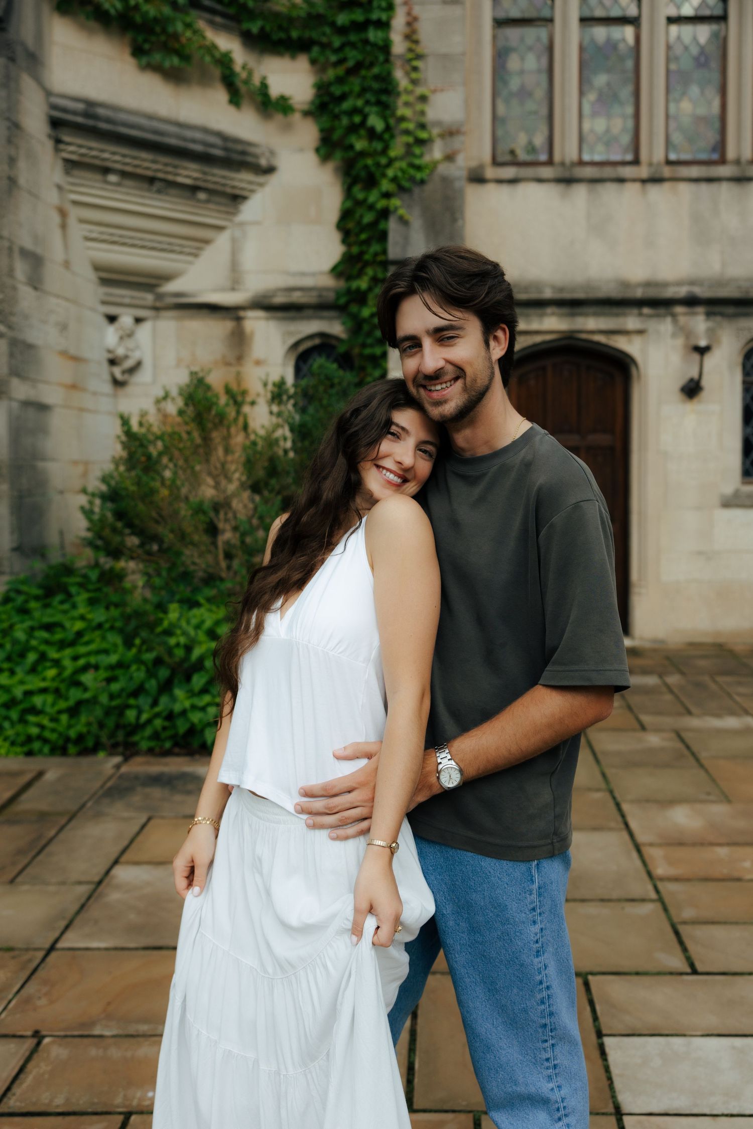 A smiling couple poses together outside a Gothic stone building covered in green ivy.