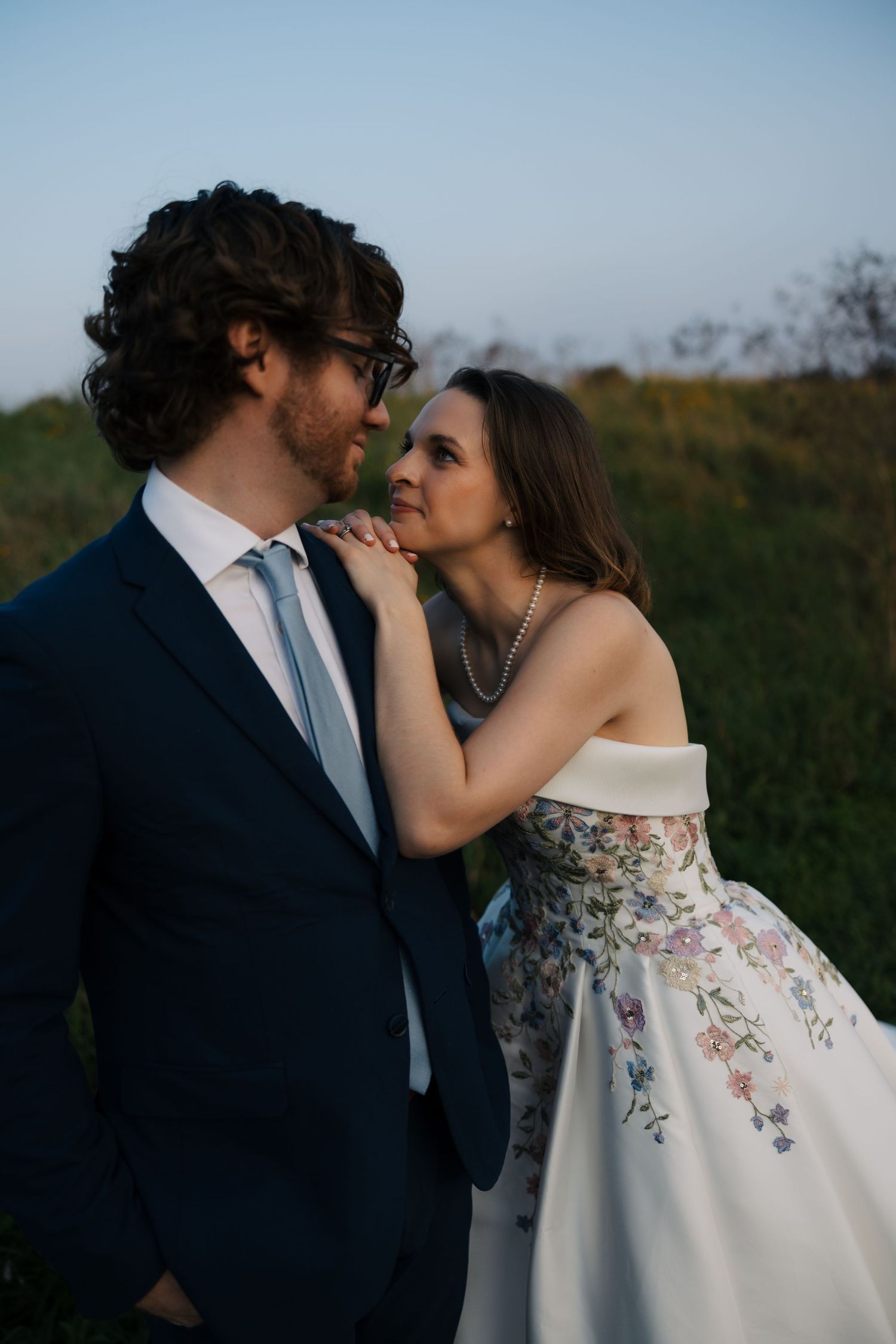 Bride in off-shoulder floral gown leans in to kiss groom in navy suit at sunset.