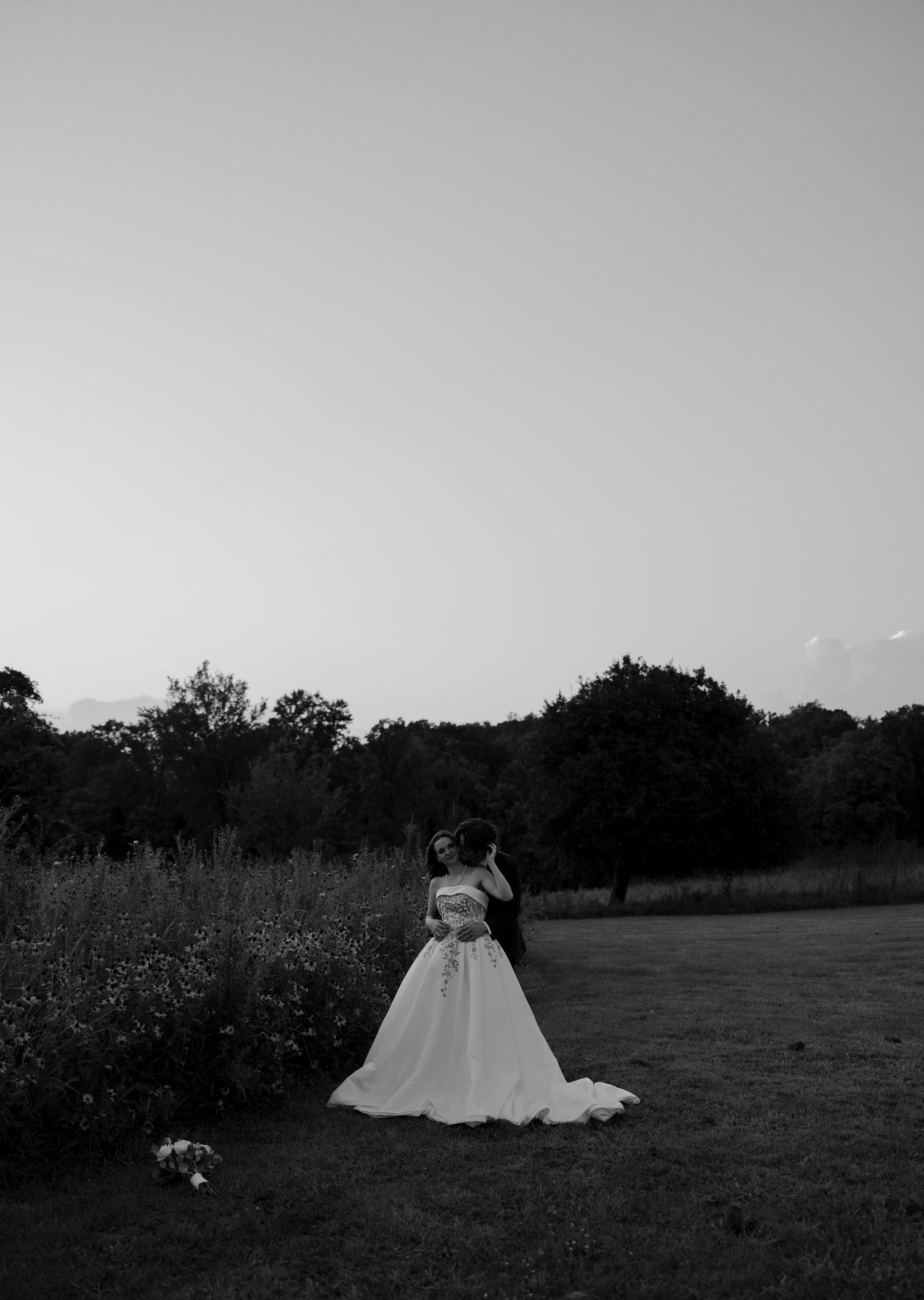 Black and white photo of bride and groom embracing in open field with trees at twilight.