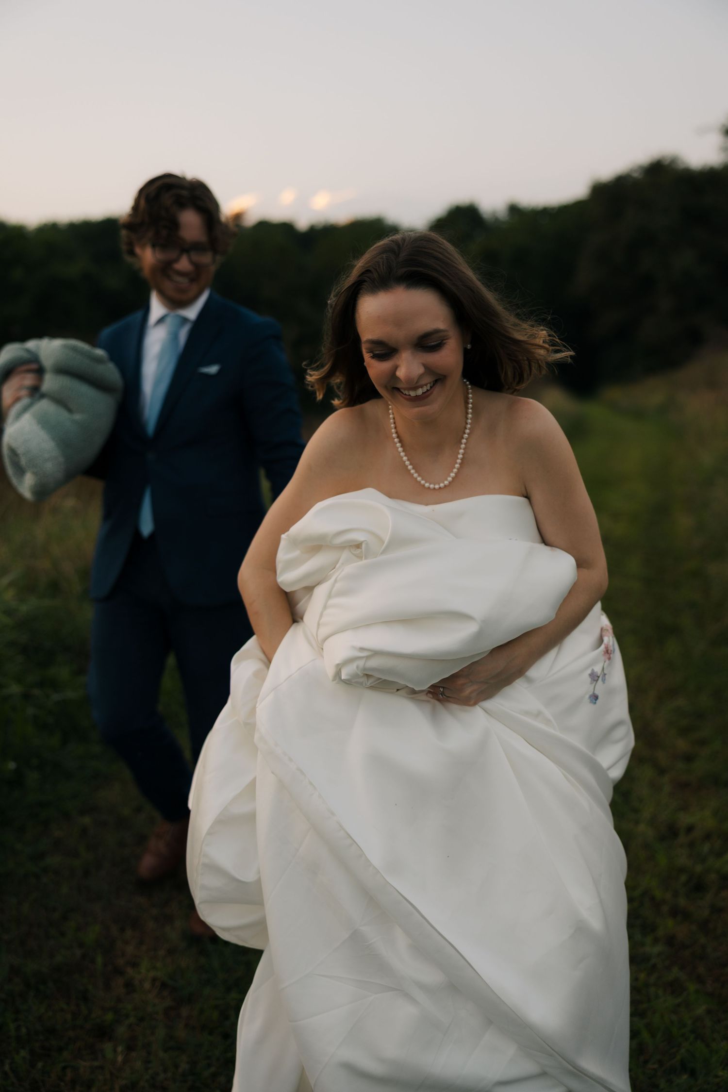 Smiling bride carries her white wedding gown train as groom in navy suit follows through a green field.