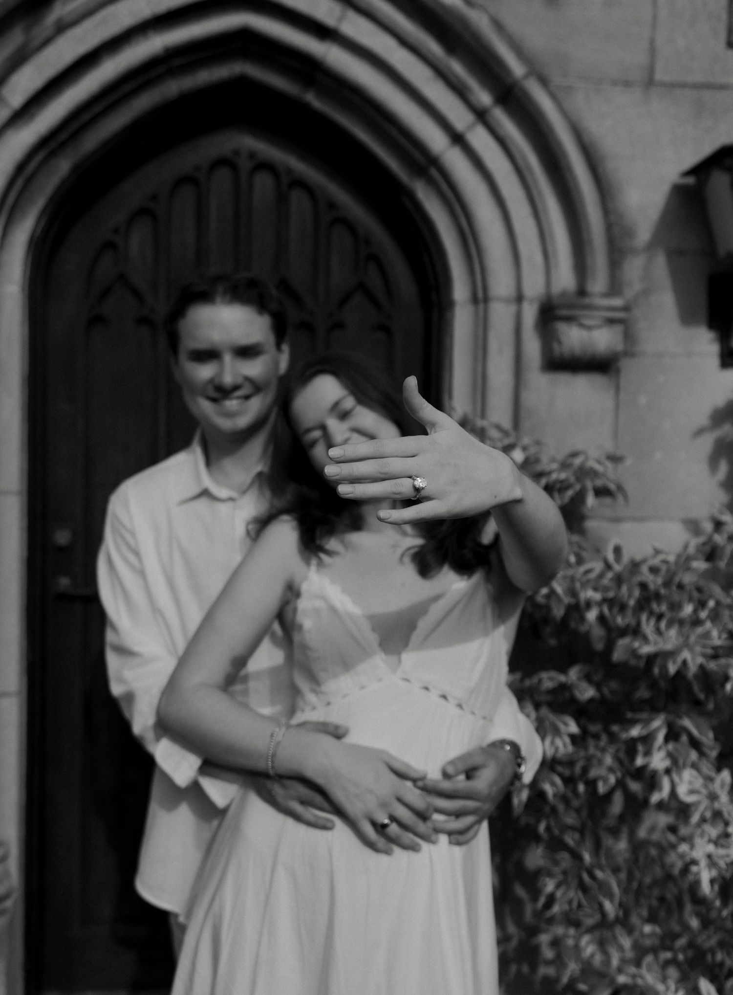 Engaged couple poses before gothic arched door as woman shows off her ring in B&W photo.