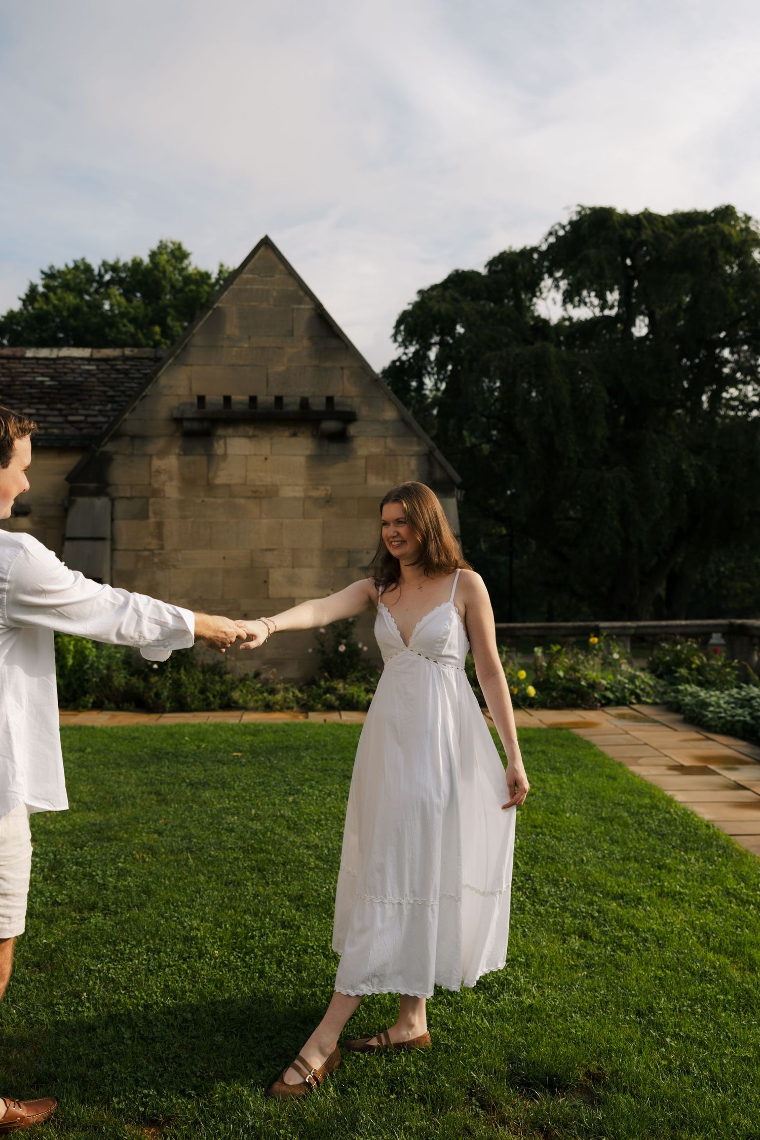 Smiling woman in white sundress holds hands with man on lawn by stone building.