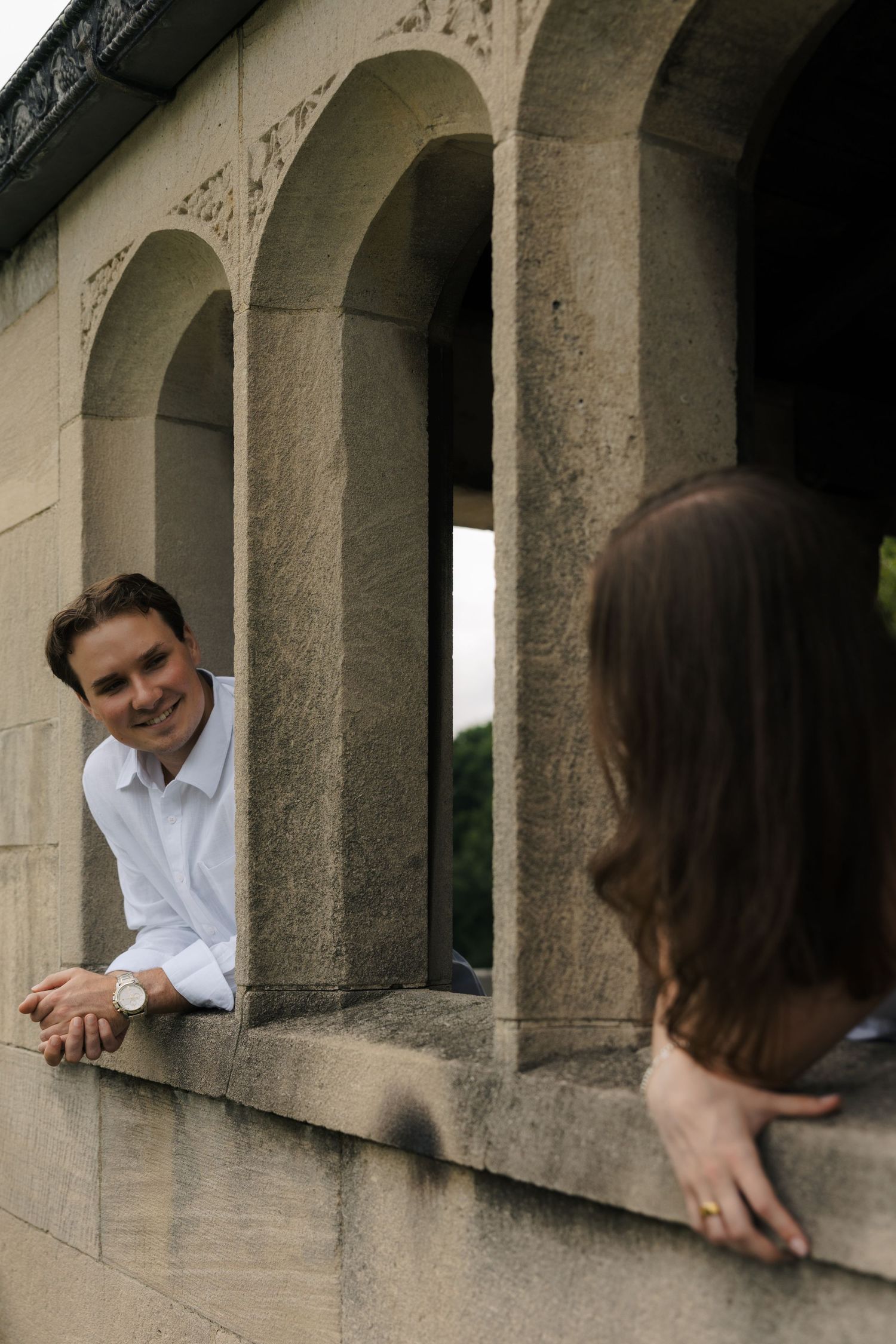 Smiling man in white shirt leaning on stone balustrade with arched windows, looking at woman in foreground.