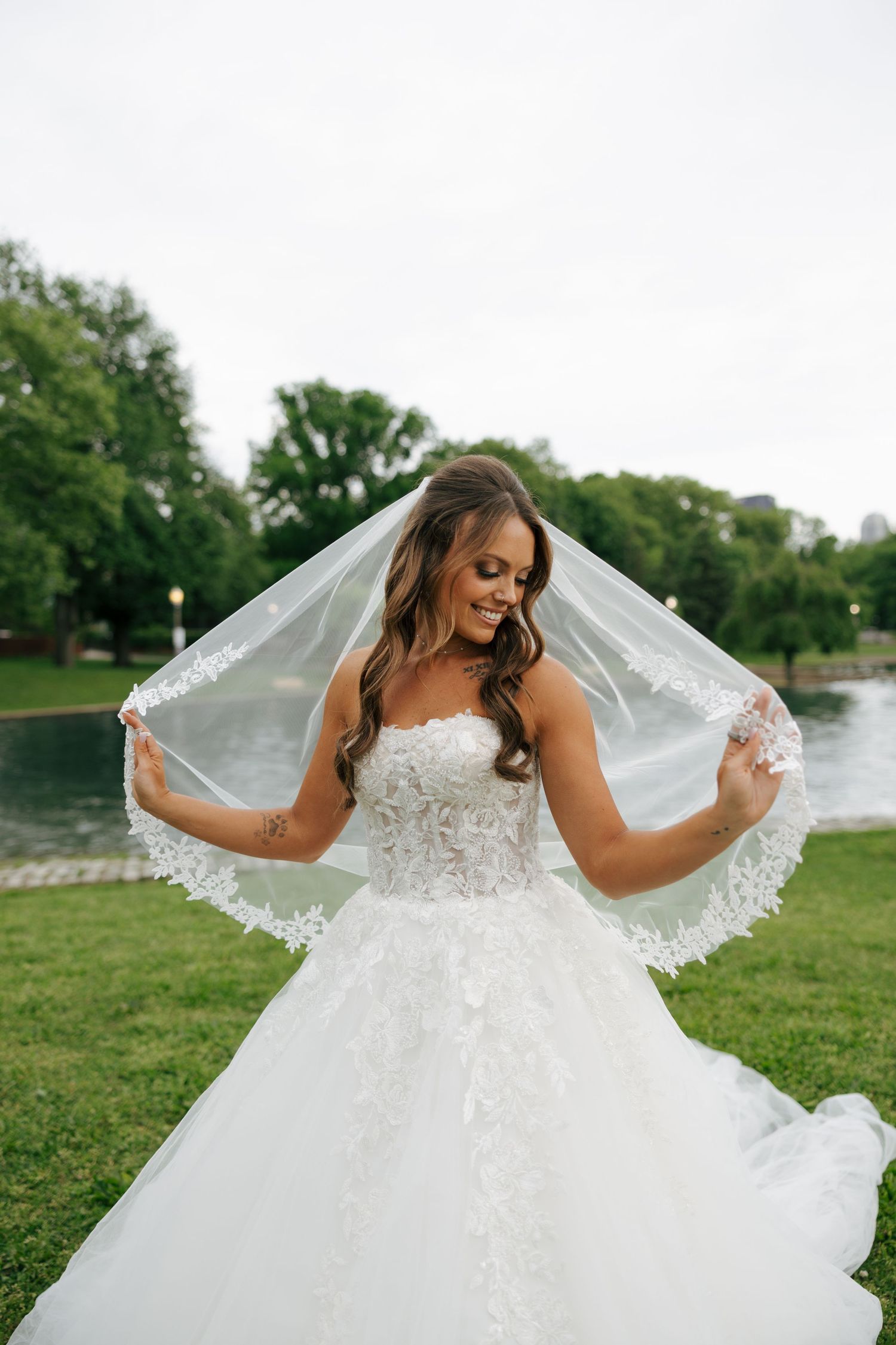 Bride in white lace ballgown holds veil wide outdoors near pond, smiling down at her dress.