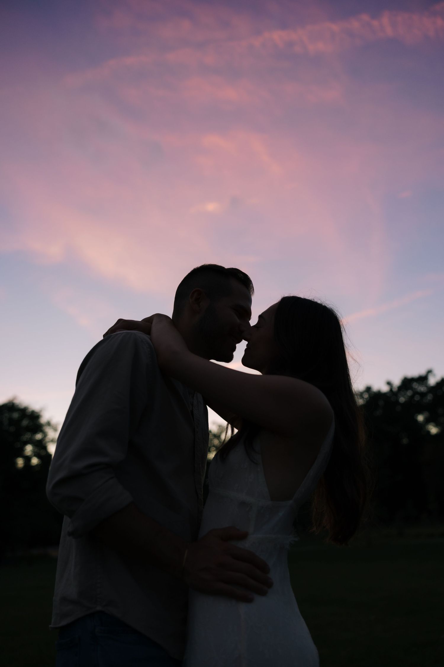 Silhouette of a romantic couple almost kissing at sunset with a pink and purple sky backdrop.