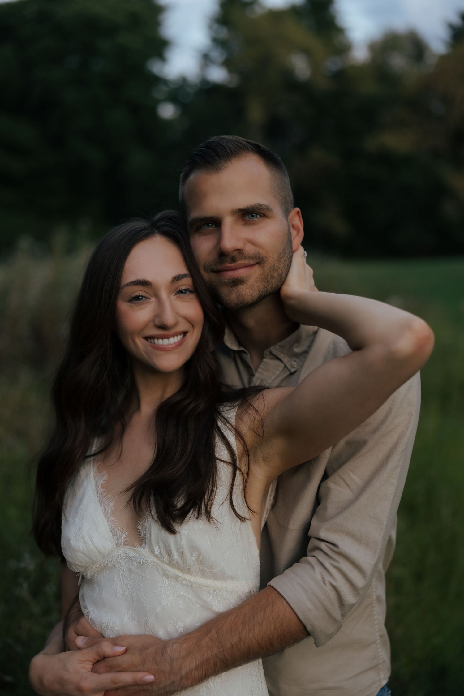 Happy couple embracing outdoors, woman in white dress smiling as man in beige shirt holds her from behind.