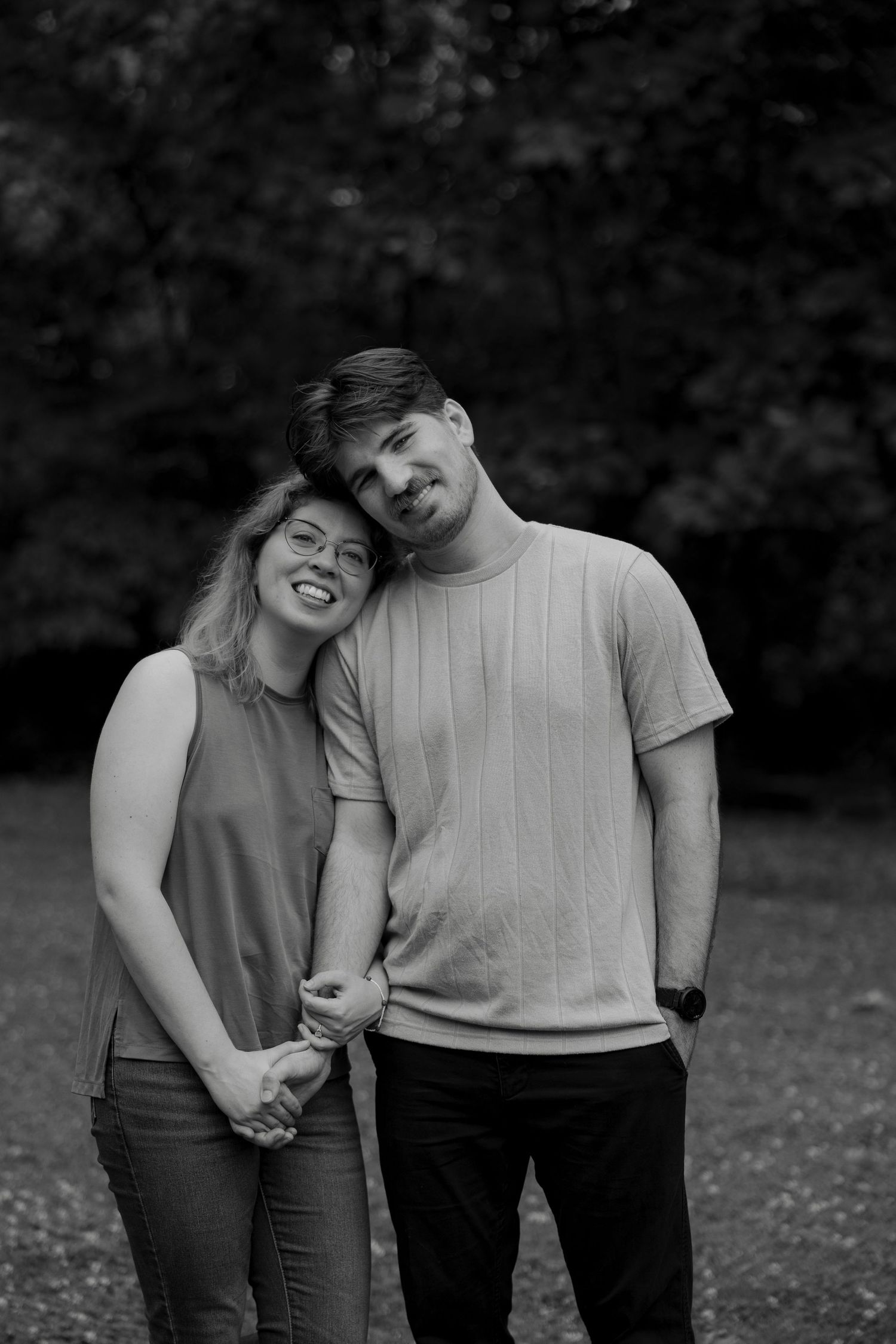 A black and white portrait of a couple holding hands and leaning together outdoors in a park.