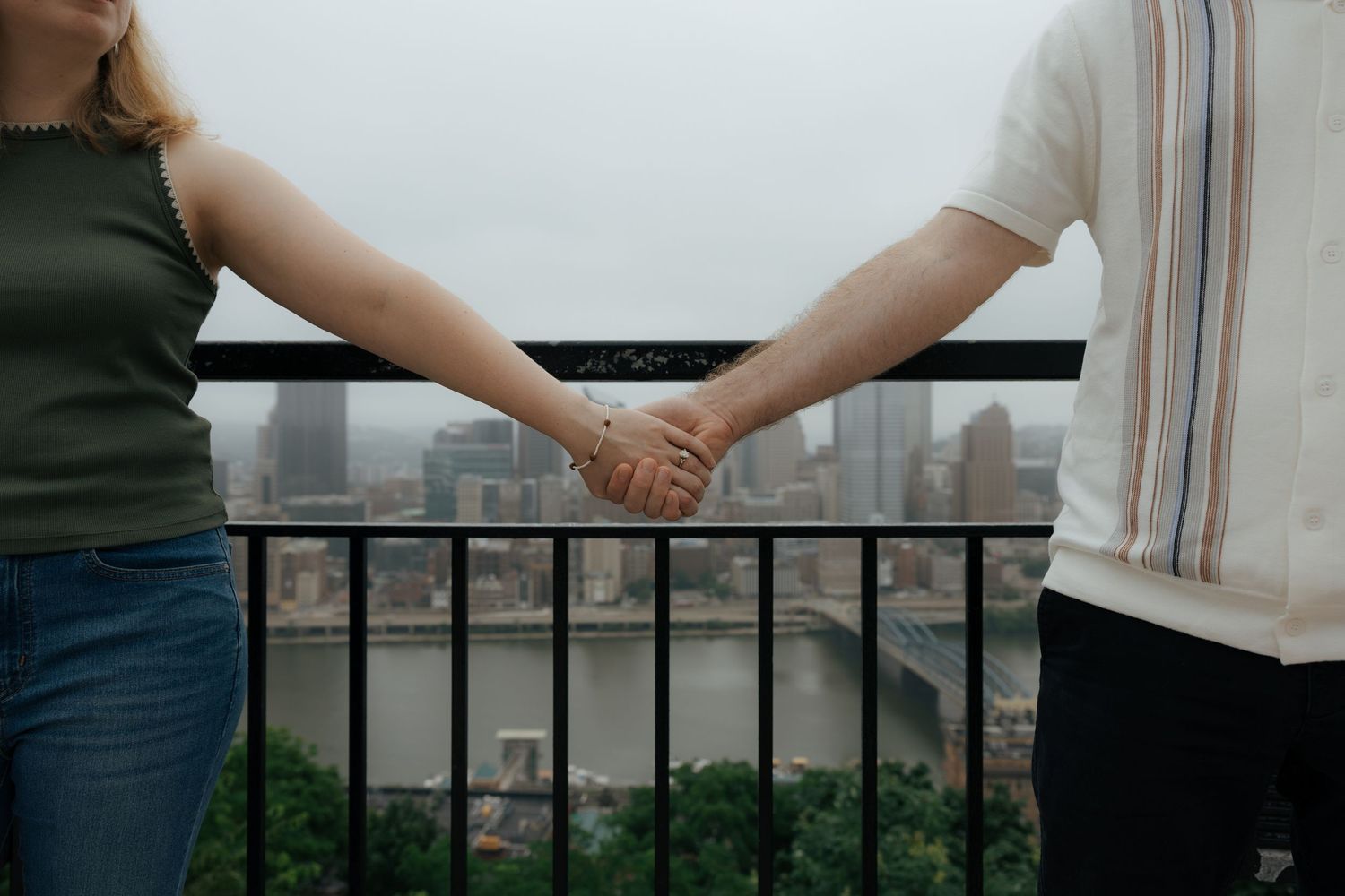 Couple holding hands on a railing overlooking a river and city bridge on a cloudy day.