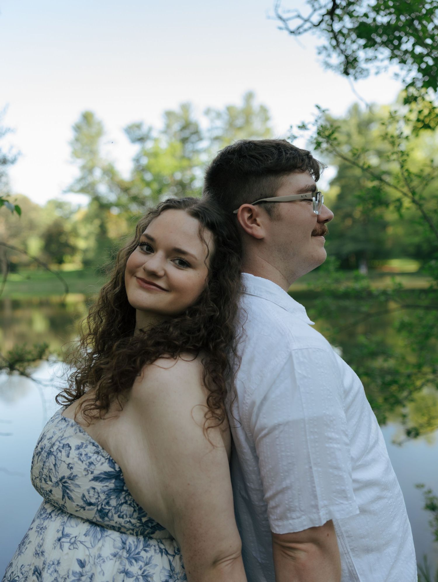 Couple posing back-to-back near a serene pond surrounded by lush green trees during a romantic outdoor photo session.